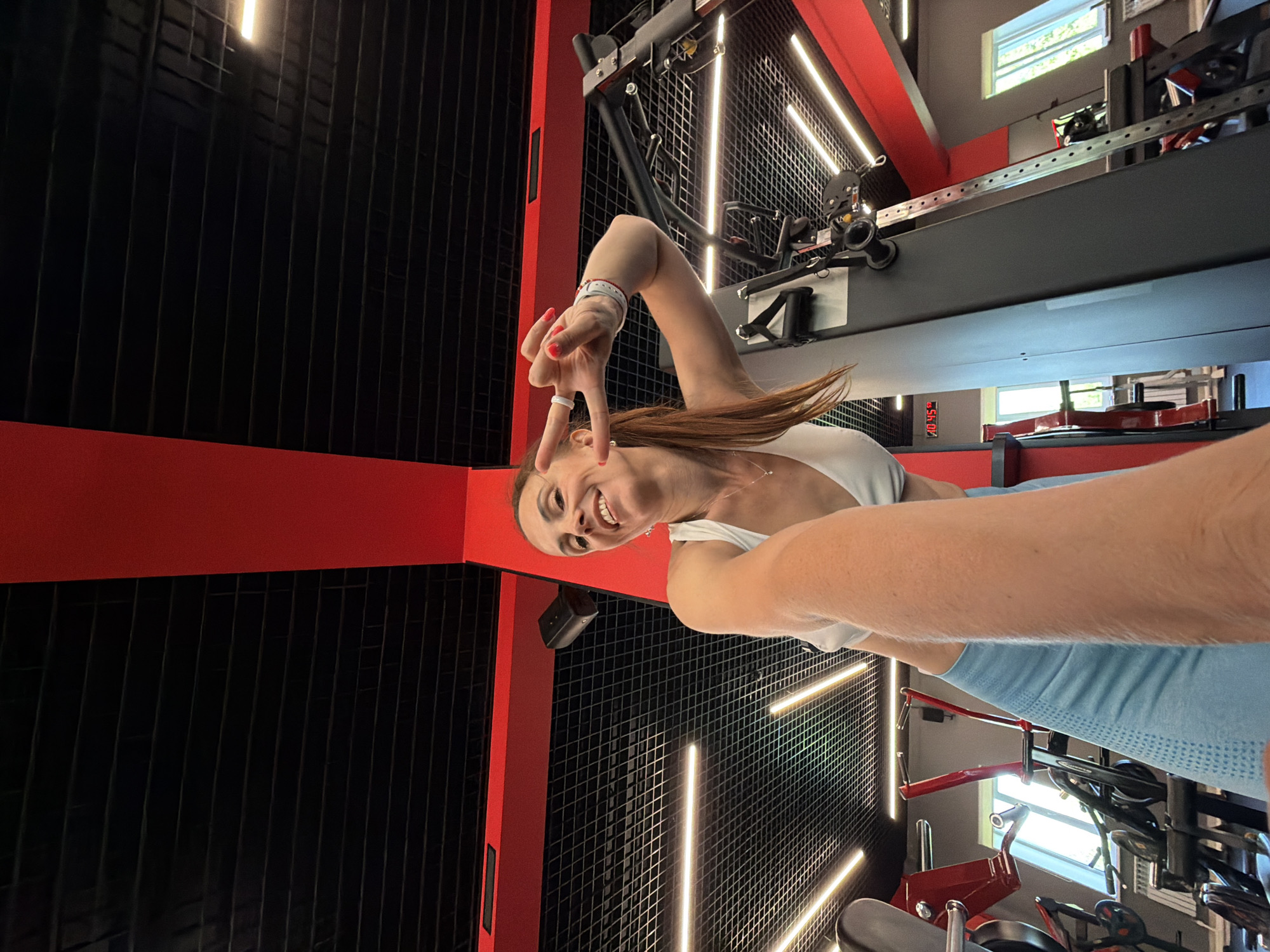 In a gym setting, a woman with her hair in a ponytail smiles and makes a peace sign with her fingers while holding a phone to take a selfie. She is wearing a white sports bra and light blue leggings. The gym features a black ceiling with red structural beams and bright fluorescent lighting. Various gym equipment, including weight machines and racks, are visible in the background. The overall environment suggests a modern and active fitness space.