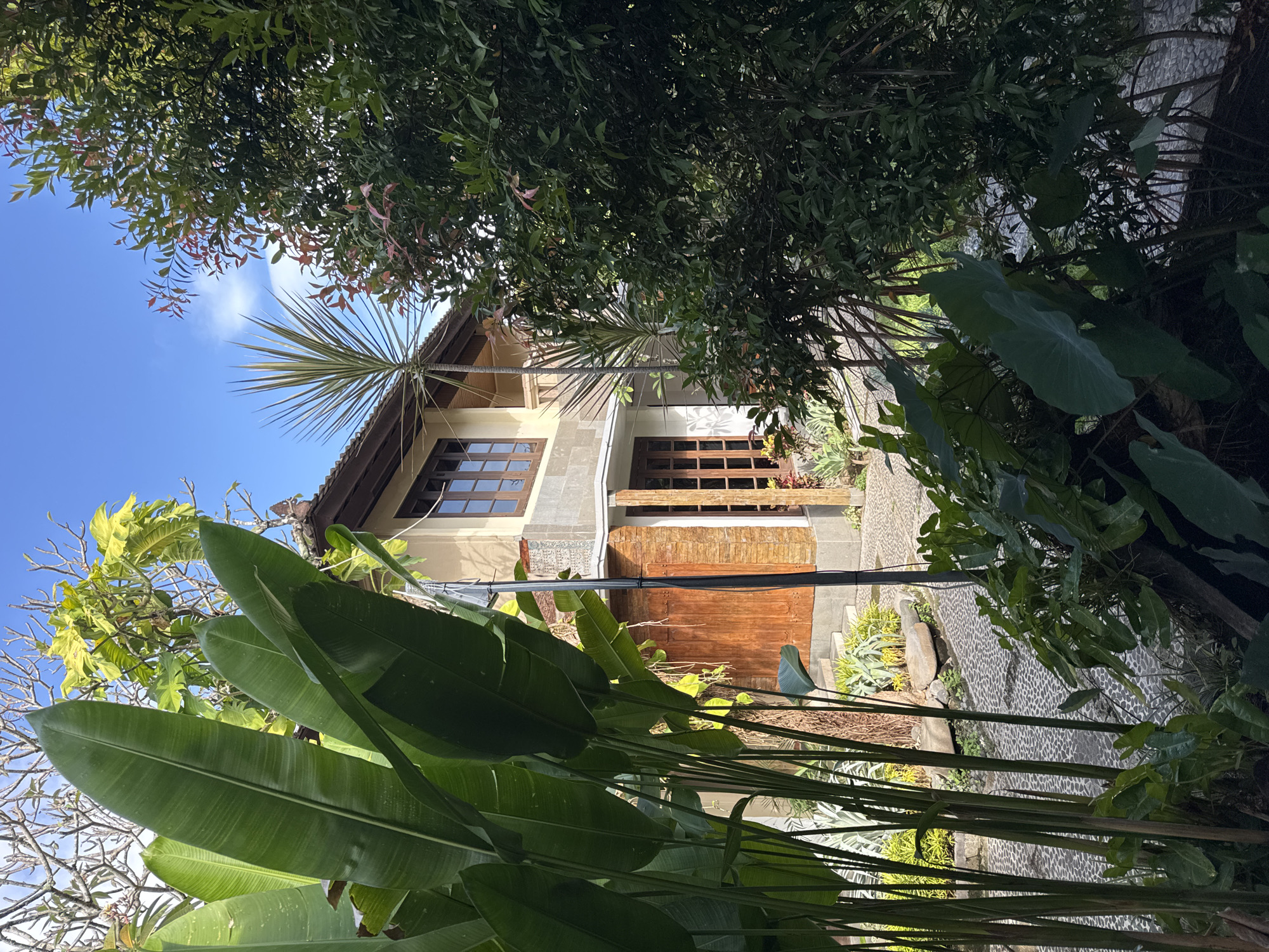 The image depicts the exterior of a building in Kuta, Indonesia, partially obscured by lush, tropical foliage. A two-story structure features a light-colored facade, dark-framed windows, and a wooden door. The entrance has a rough-hewn, wood-and-stone design. Overhanging eaves are visible beneath a cloudy blue sky, partially covered by the branches of a tall tree. The foreground includes banana leaves and various other plants. A stone-paved pathway runs alongside the building. An overhead power line is visible.
