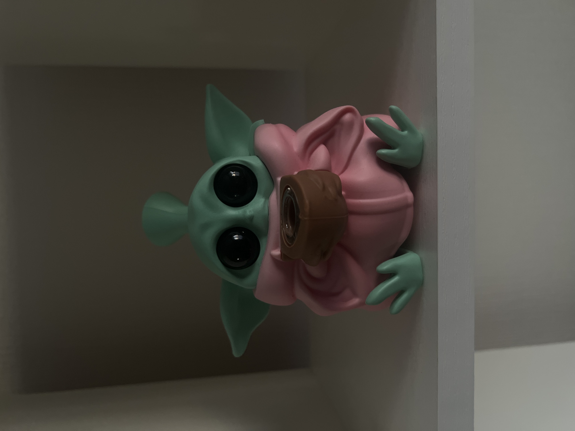 A small, green figurine resembling the character Grogu from Star Wars is positioned centrally on a white shelf. The figurine has large, black eyes, pointed ears, and is wearing a pink robe. It is holding a brown cup in its hands. The background consists of a dark, muted wall. The lighting is dim, casting shadows on the figurine and the shelf. The image was taken in Saint Petersburg, Russia.