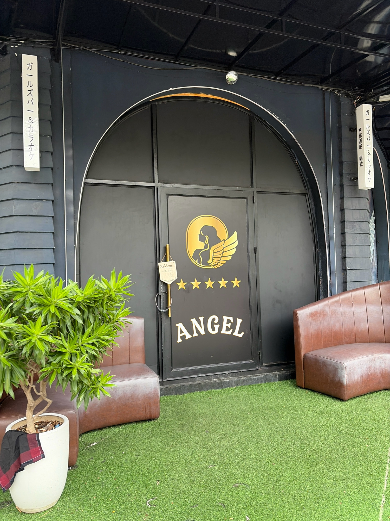 The image depicts the entrance to a business in Da Nang, Vietnam. The entrance features a large arched black door with a gold logo of a woman with wings and the word "ANGEL" in gold lettering. A small "Welcome" sign is hanging from the door handle. To the left of the door is a green potted plant, and to the right are two brown leather bench seats. Artificial grass covers the ground in front of the entrance. Two vertical signs, one on each side of the entrance, display Japanese text. The overall impression is that of an entertainment venue.