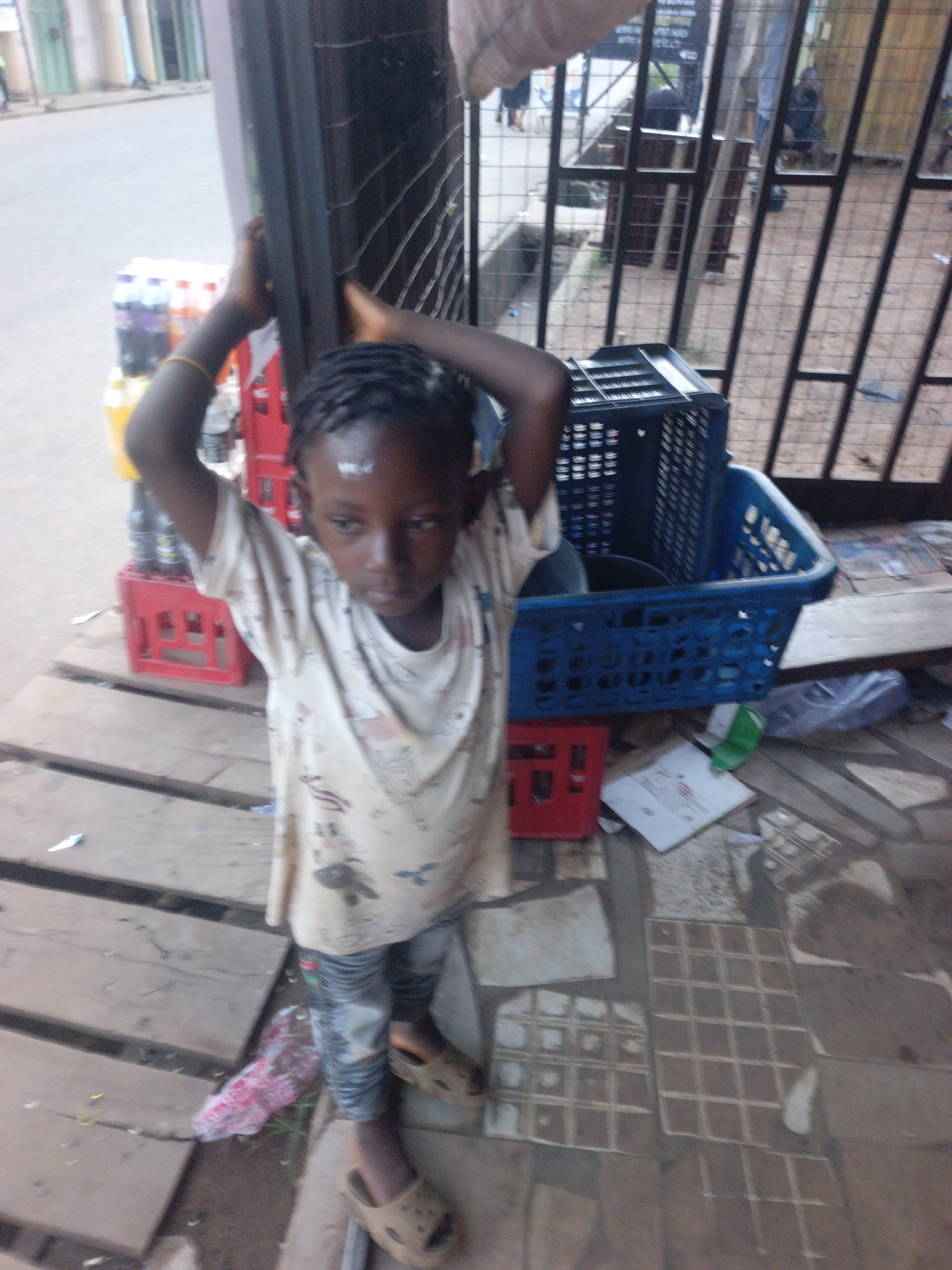 A young, dark-skinned child with braided hair, wearing a light-colored, patterned t-shirt, mottled jeans, and sandals, stands with arms raised above their head, holding onto a dark-colored, vertically lined frame or structure. The child appears to be on a sidewalk or porch constructed of wood planks and mismatched tile sections. Behind the child are red crates containing bottled drinks, a blue plastic basket containing what appears to be a black pot, and a black crate. Debris and litter are scattered around the immediate area, including papers and plastic wrappers. In the background, through a metal barred fence, there is a glimpse of a street and a few figures. The scene appears to be in Akwanga, Nigeria.