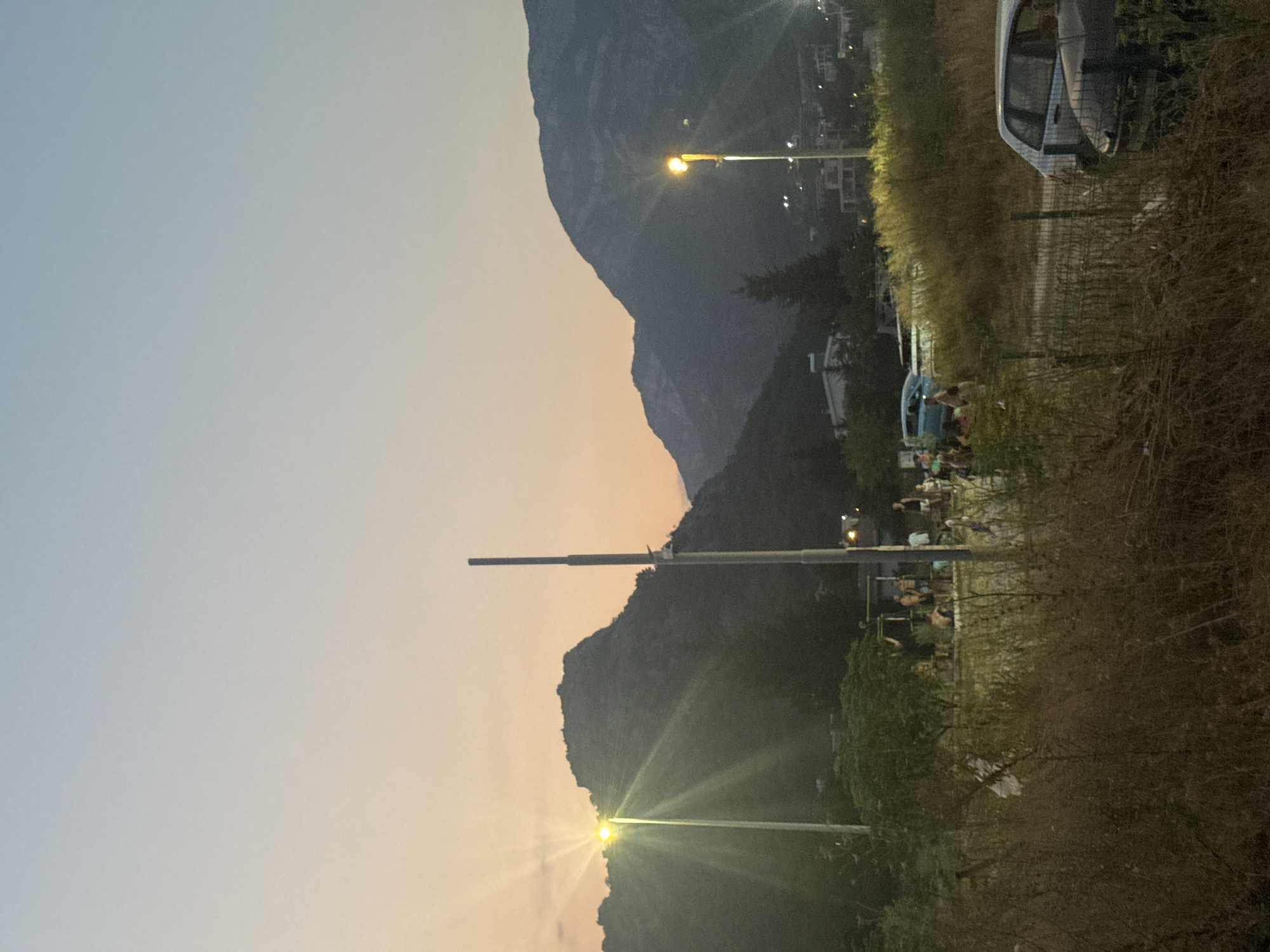 At dusk, in the city of Sutomore, Montenegro, a group of people gather in a field of dry grass and brush. In the background, silhouetted mountains rise against a gradient sky transitioning from pale orange near the horizon to a soft blue. Two prominent streetlights illuminate the scene, one on the left casting a strong, starburst-like glare, and another on the right with a softer glow.
A group of approximately ten individuals are visible in the mid-ground, some standing, others appearing to be seated or interacting. Their clothing suggests a casual, warm-weather setting. Several cars are parked in the area; a light-colored car is positioned on the right side of the frame, and a darker car is partially visible in the foreground. A blue van is also present amongst the group. Scattered vegetation, including trees and tall, dry grasses, occupies the foreground and mid-ground. A tall, thin pole, possibly a lamp post or utility pole, stands centrally in the mid-ground. The overall impression is a casual outdoor gathering in a semi-rural or outskirts area of the city as daylight fades.