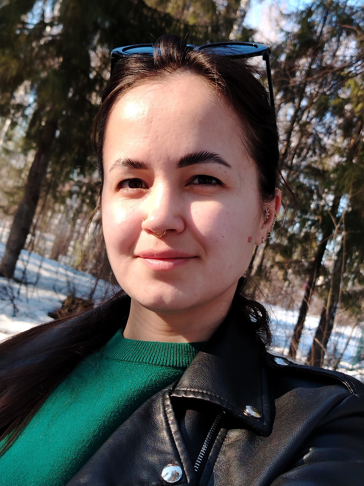 A woman is taking a selfie outdoors in what appears to be a wooded area. She is wearing a green sweater under a black leather jacket. Her dark hair is pulled back. She has a nose ring and multiple earrings in her left ear. Sunglasses are perched on top of her head. The background shows trees with snow on the ground. The city of Noviy, Russia, is not apparent from the provided information.