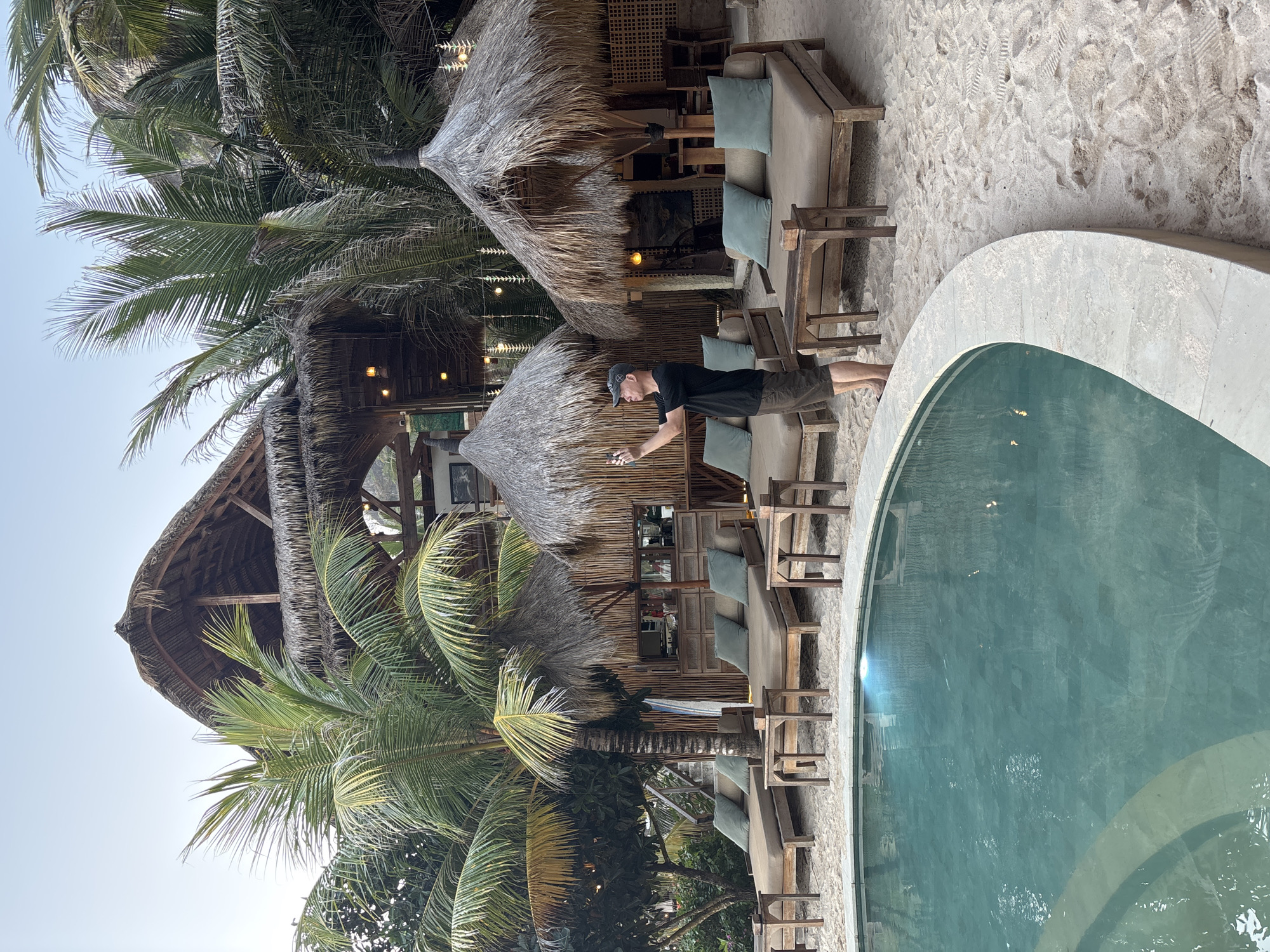 A man in a dark t-shirt and shorts stands on a sandy beach in Kuta Selatan, Indonesia, holding a smartphone and taking a photo of a multi-level, open-air thatched-roof building. In the foreground, a curved swimming pool with turquoise water is visible. The building features multiple levels, with thatched roofs and bamboo walls. Several lounge chairs with light-colored cushions are arranged on the sand in front of the building. Palm trees surround the area. The scene appears to be a resort or beach club.