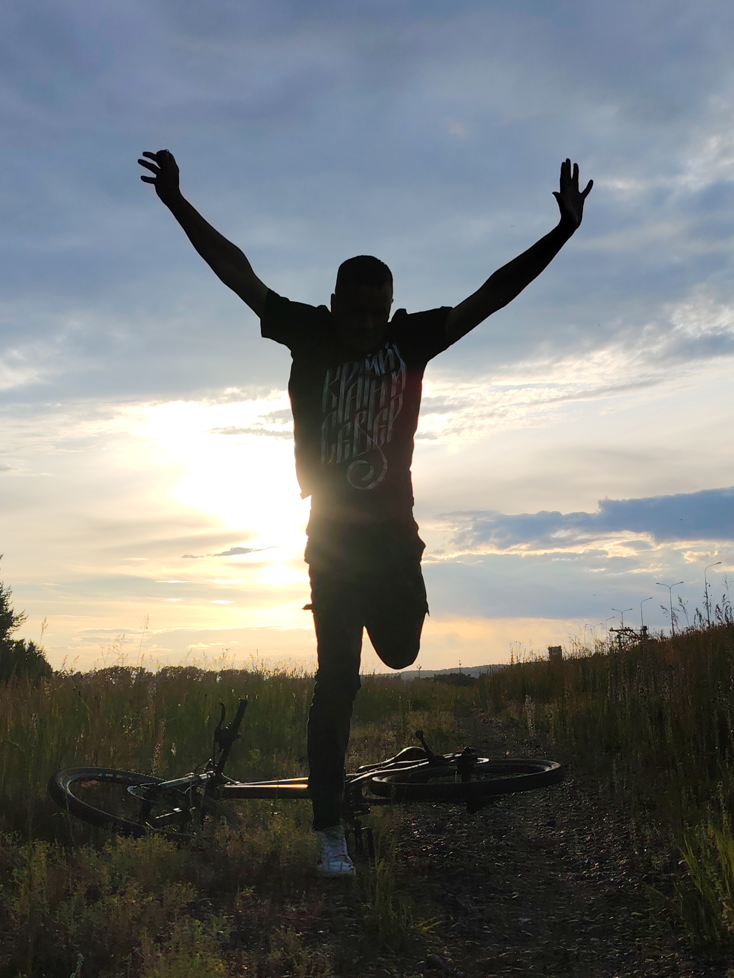 A silhouette of a person stands in a field at sunset, with their arms outstretched towards a cloudy sky. The sun is low on the horizon, casting a warm glow and creating a lens flare effect. A bicycle lies on its side in the foreground, partially obscured by tall grass and weeds. The ground is a mix of dirt and gravel. In the background, trees can be seen on the left, and a line of streetlights runs along a slightly elevated area on the right. The scene evokes a sense of freedom and accomplishment, possibly after a cycling adventure. The person is wearing dark clothing, including a t-shirt with visible text. The text on the shirt appears to be in a stylized font and reads "BRAVE HEART". The weather is cloudy with hints of blue sky peeking through. The overall mood is serene and optimistic.