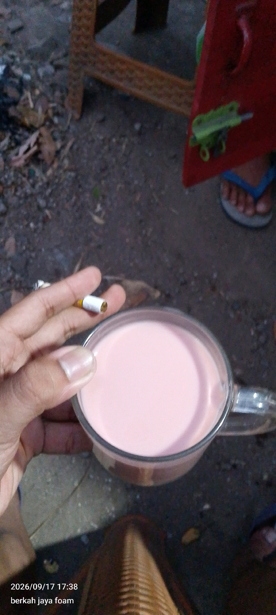 A close-up, low-angle shot shows a hand holding a cigarette over a glass of pink liquid. The background consists of a textured surface resembling dirt or gravel, with some scattered leaves and debris. To the right, a red object, possibly a piece of furniture or equipment, is partially visible, with a small green plastic object attached to it. A person's foot wearing a blue flip-flop is also in the frame, near the red object. In the bottom left corner, text overlay reads "2026/09/17 17:38 berkah jaya foam". The scene appears to be outdoors, possibly in an industrial or utility area, given the debris and the rough texture of the ground. The lighting suggests it is late afternoon or early evening. The image does not provide specific landmarks to pinpoint the exact location within Kraton, Indonesia.