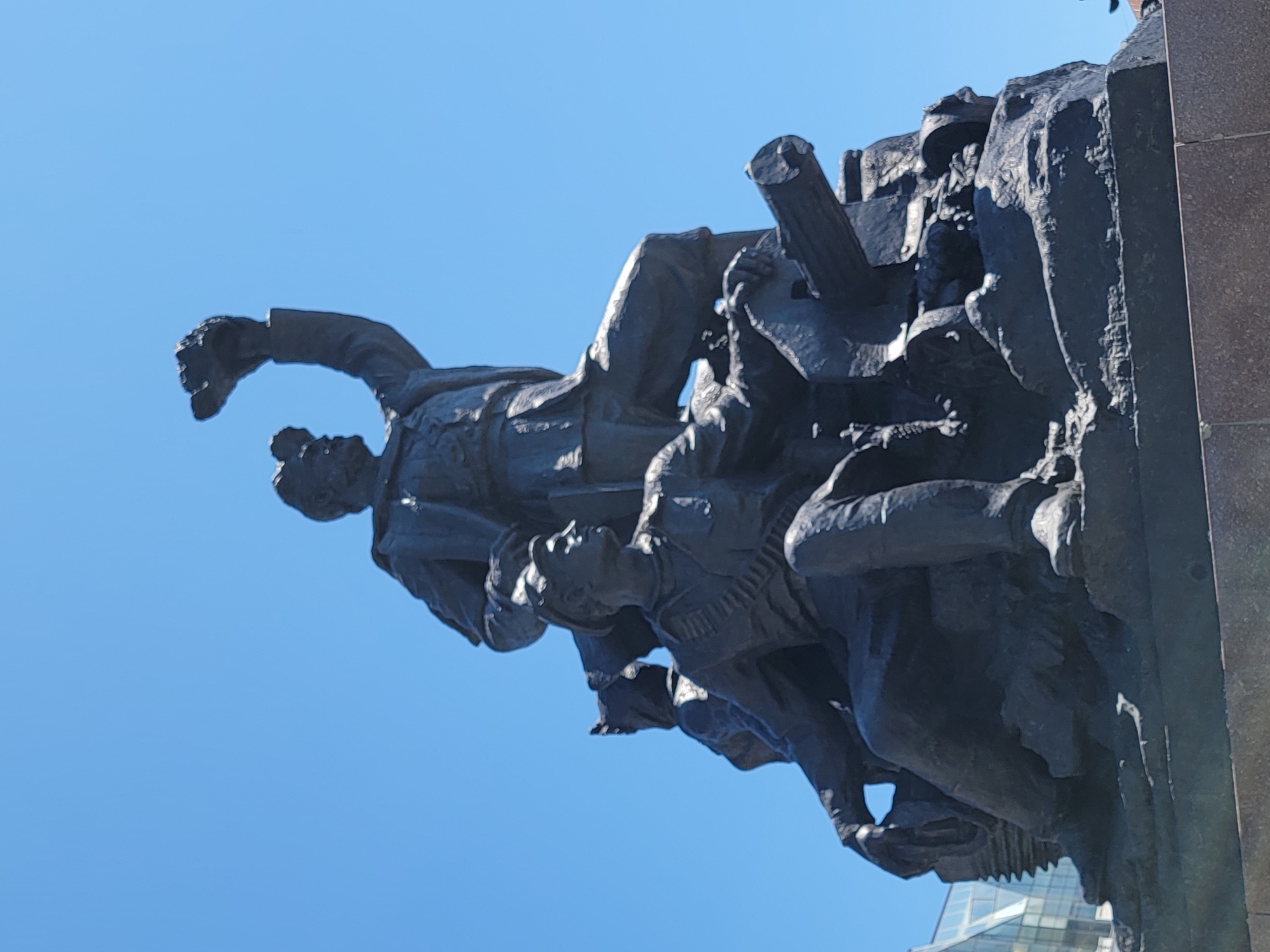 The image captures a low-angle view of a dark, possibly bronze, sculptural monument set against a clear blue sky. The sculpture depicts a group of figures in dynamic poses, possibly representing soldiers or laborers engaged in a struggle or heroic action. The figures are clustered together, with one figure prominently raising his fist. The monument is located in an urban environment. Based on the architecture visible in the lower right of the image, with a modern glass building, it is likely to be in a major city. The location in the city of Unknown, Unknown is unconfirmed.