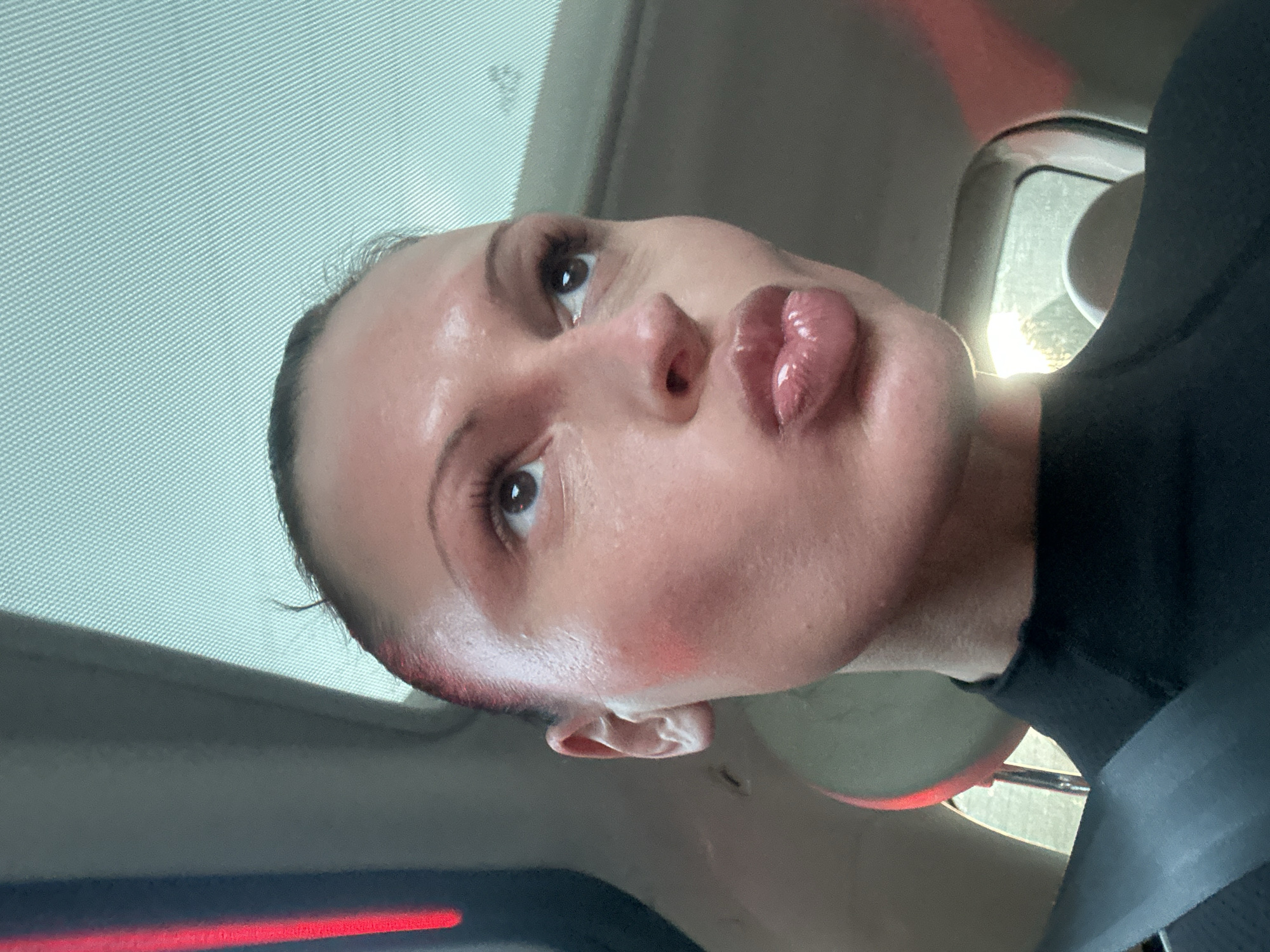 A female individual is depicted in a vehicle interior, seen from the chest up. She wears a dark, high-necked garment and a grey seatbelt across her left shoulder. Her hair is pulled back, revealing a clear forehead. Her face is turned slightly to the right, with eyes gazing upwards and to the right. Her lips are pursed in a pouting expression, appearing glossy. Pink blush is visible on her cheeks. Overhead, a textured, light-colored sunroof screen is visible. To the left, a dark car pillar is present with a vertical red light reflection. Bright exterior light enters from the lower right, highlighting part of the vehicle's interior. No visual information within the image identifies the location as Tunbridge Wells, United Kingdom, or any specific point within it.