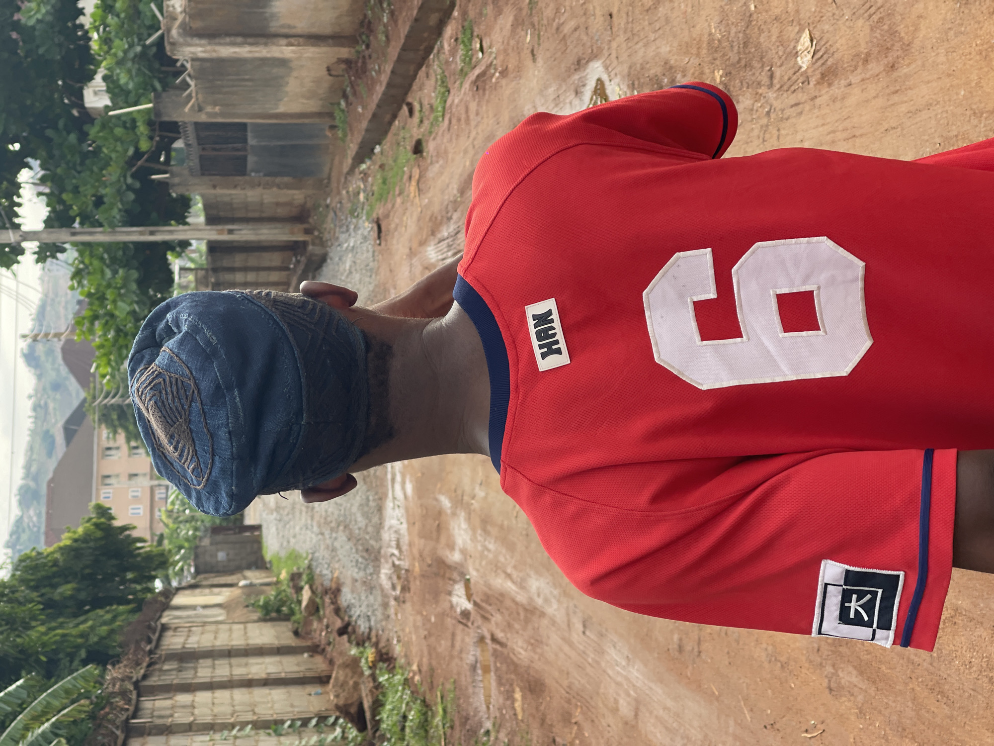 A person wearing a blue cap and a red jersey with the number 6 on the back is standing on a dirt road in Dutsen Alhaji, Nigeria. The jersey also has a tag labeled "HAN" near the collar and a square logo with a "K" on the sleeve. The road appears to be unpaved and slightly muddy. Buildings and vegetation are visible in the background.
