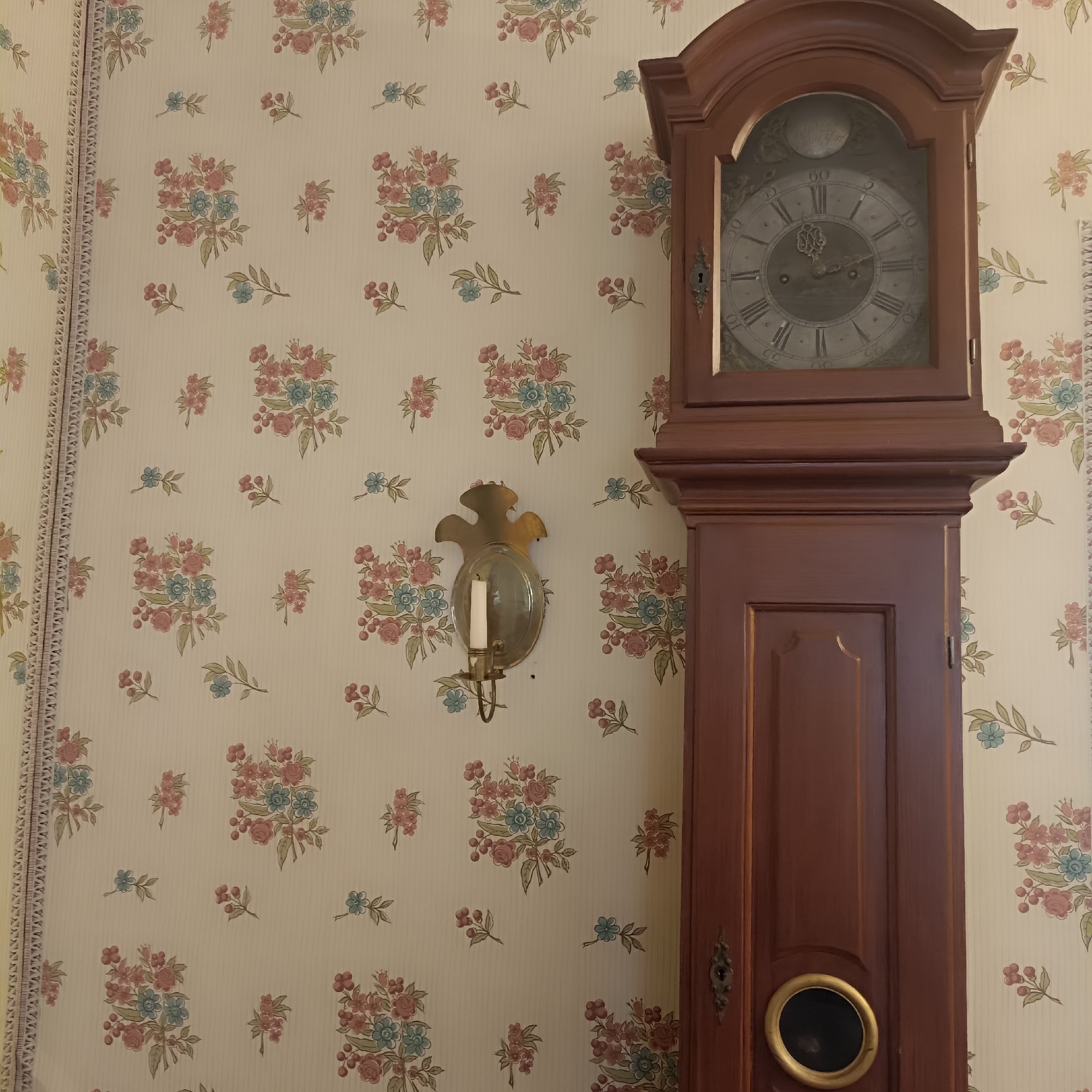 The image shows a close-up view of a grandfather clock and a wall sconce against floral wallpaper. The grandfather clock is made of dark wood and has a circular glass face displaying Roman numerals. The wall sconce is made of brass and holds a single white candle. The wallpaper has a cream background with a pattern of small bouquets of pink and blue flowers. A decorative trim runs along the left edge of the wallpaper. The scene is set indoors, in what appears to be a home or a museum. There is no information available to determine the specific location in the city of Frankfurt, Germany.