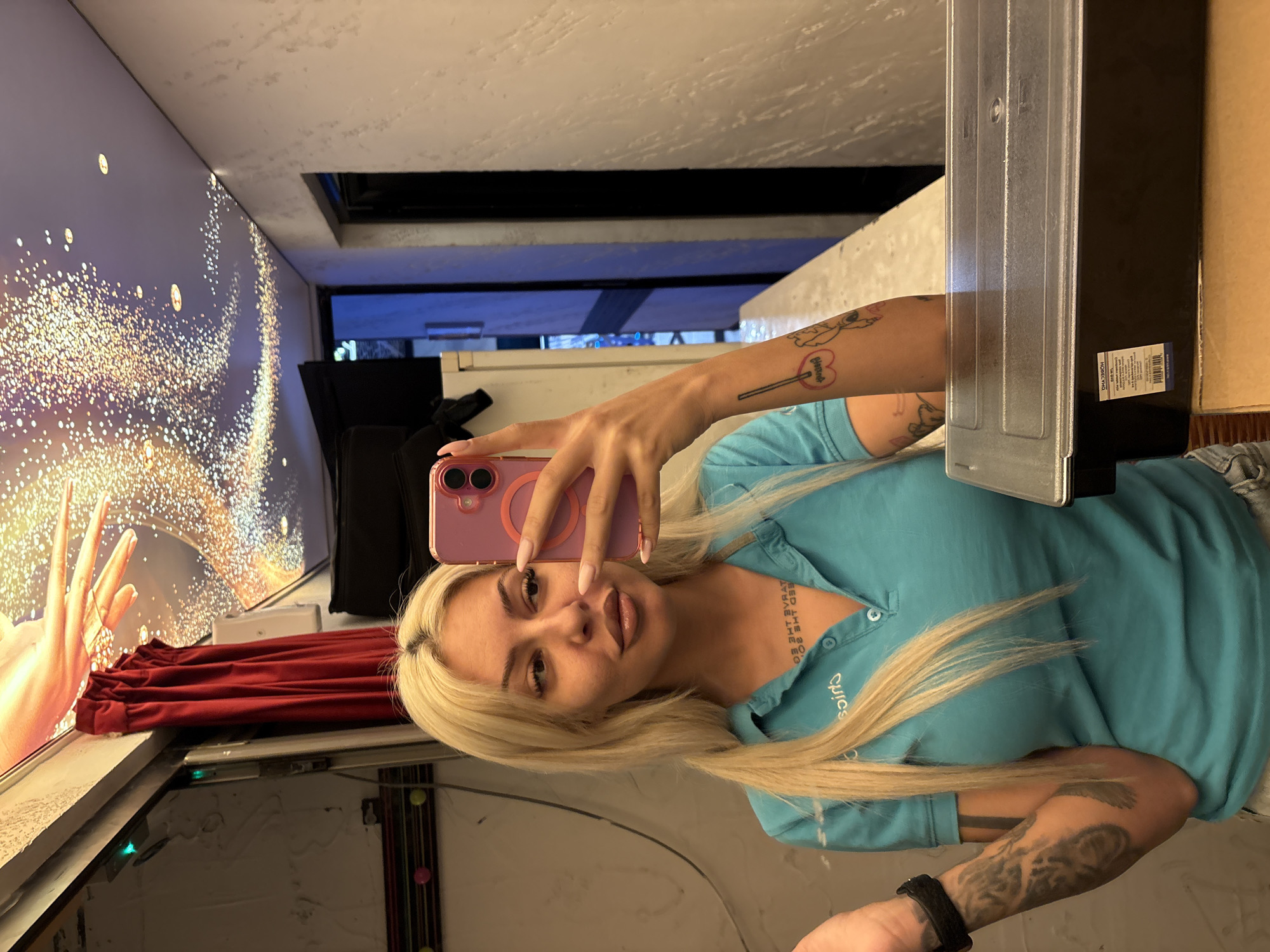 A woman with blonde hair is taking a selfie indoors, holding a pink smartphone. She wears a light blue collared shirt, and has visible tattoos on her neck and arms. Behind her, there is a window displaying what appears to be a projected image of light patterns and what might be part of a hand. A red curtain or drape is also visible near the window. The location may be Plovdiv, Bulgaria, but there is no immediately discernible sign of this within the provided image. A black object is on the right side of the image.