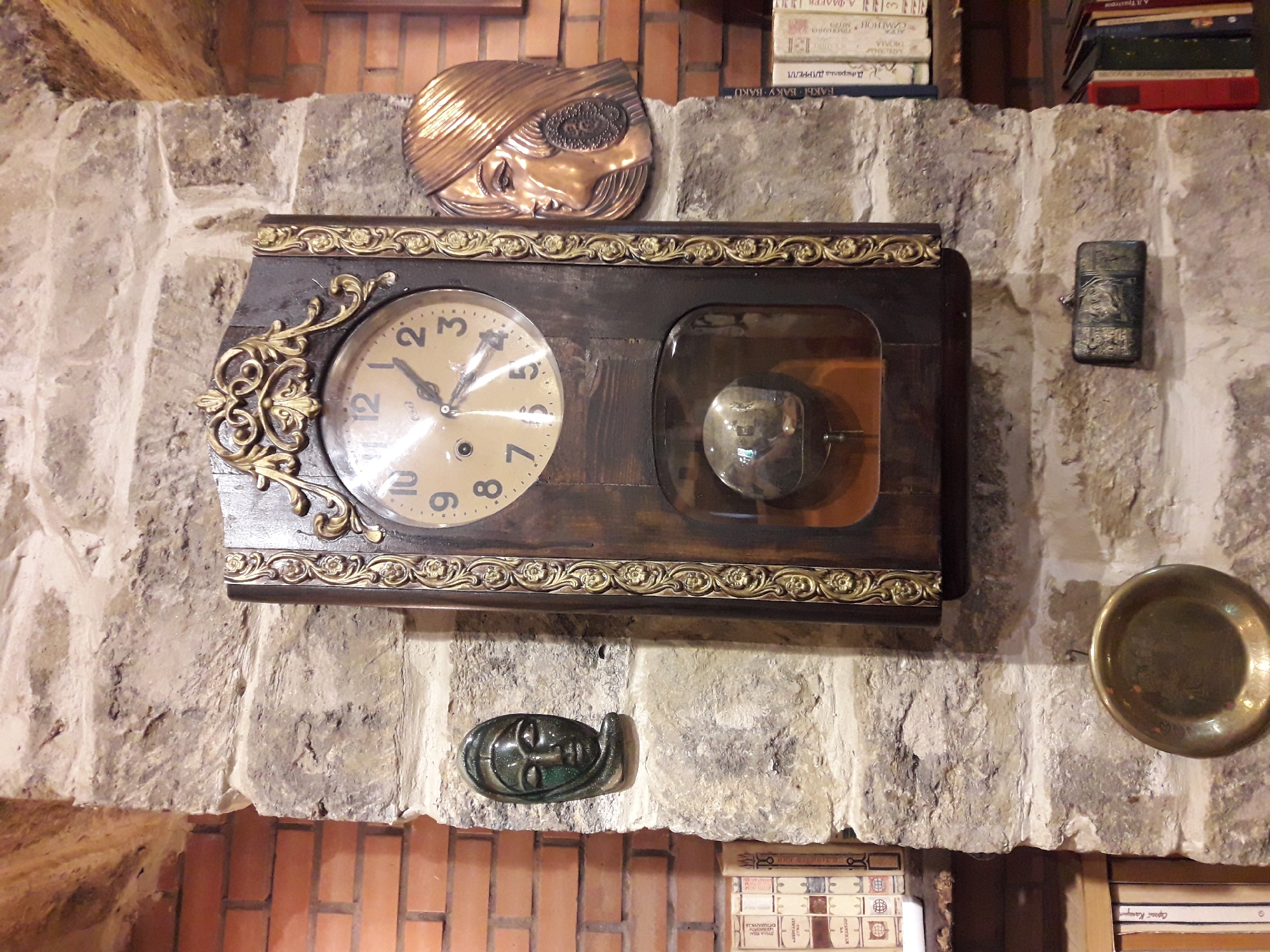 An antique pendulum clock hangs on a stone wall, flanked by brickwork. To the left of the clock is a small, dark green ceramic mask of a face with closed eyes. To the right of the clock is a copper relief of a woman's face in profile, with a large circular earring. Below the clock, a small, dark metal box is attached to the wall. Further down, a brass plate hangs from a hook. Bookshelves filled with books are visible on the left and right sides of the image, suggesting a study or library setting. The clock's face shows Roman numerals and has two hands indicating a time just past 12:30. The pendulum is visible through a glass pane at the bottom of the clock. The overall impression is one of an eclectic collection of decorative items and books displayed in a rustic environment.