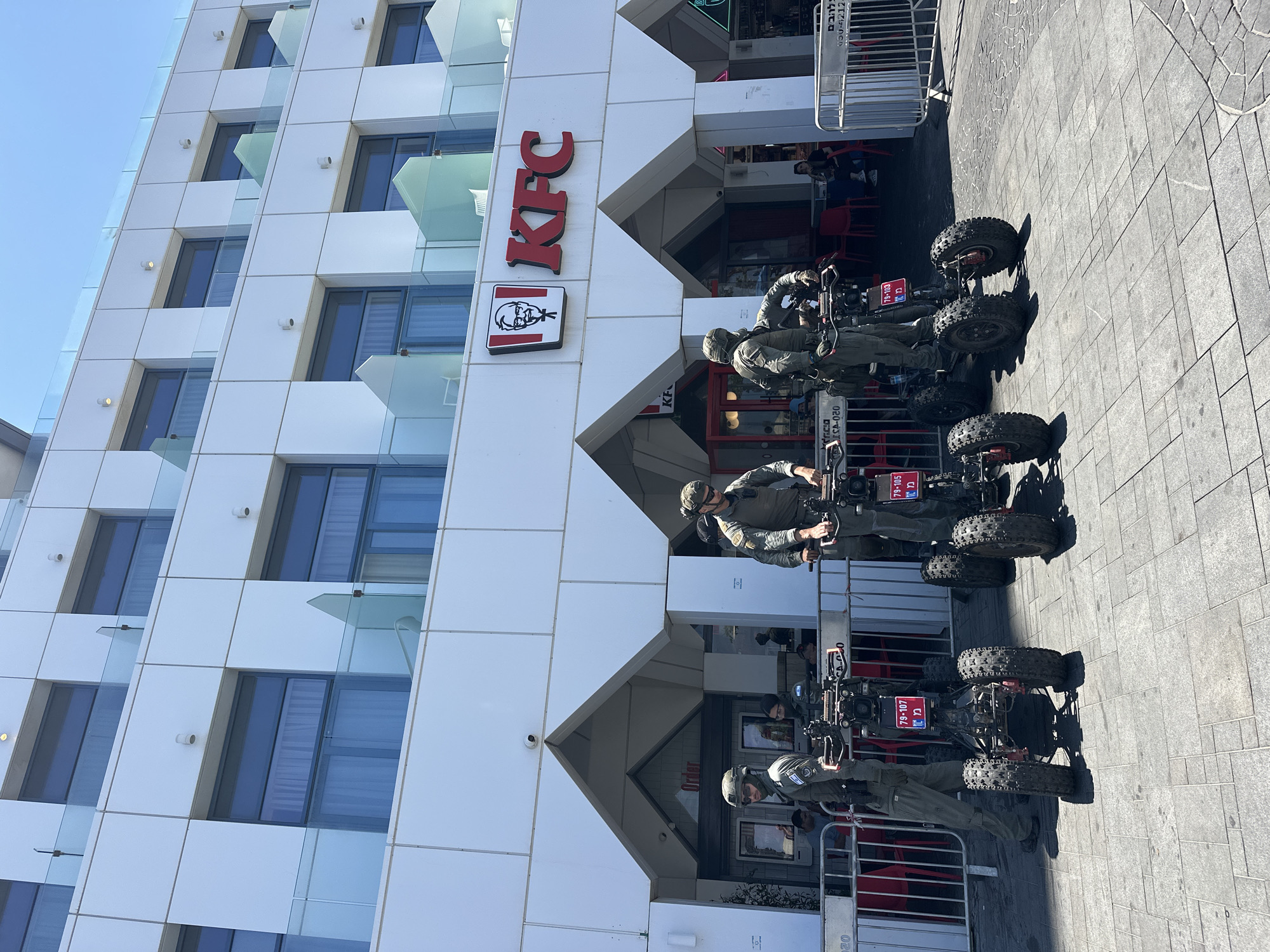 In Tel Aviv, Israel, a bright sunny day is captured with a modern white building facade. The building has multiple levels of windows, some with blue curtains, and glass balconies. The prominent red and white KFC logo is displayed on the upper level. Below, at ground level, four individuals in military-style uniforms are positioned on quad bikes. The individuals are wearing helmets and tactical gear, with two of them standing on the quad bikes which are equipped with large, knobby tires. Red and white metal barriers are set up in front of the KFC entrance, with some people visible sitting at tables inside. The ground is paved with grey square tiles. The scene appears to be a public area outside a fast-food restaurant.