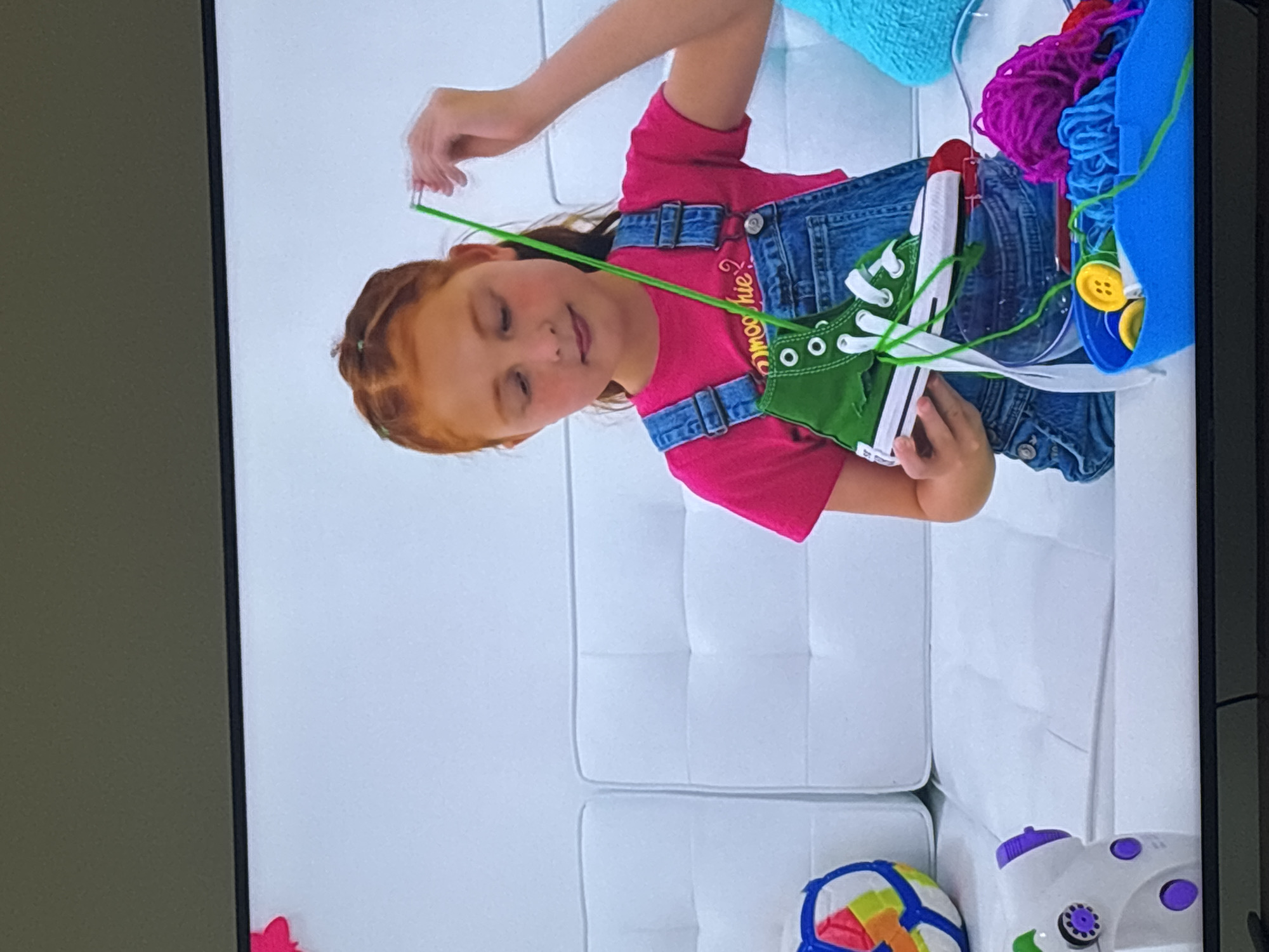 A young girl with red hair, wearing a pink t-shirt and denim overalls, sits on a white tufted couch. She is holding a green high-top sneaker with white laces and is threading green yarn through the eyelets. In front of her is a blue plastic container filled with yarn of various colors, including purple, blue, and red, along with yellow buttons. To her left, a colorful ball and a white toy sewing machine with purple accents are visible on the couch. The scene appears to be indoors, with a plain white wall behind the couch. The image is a screenshot from a digital display. There is no indication of the location being Dubai, United Arab Emirates.
