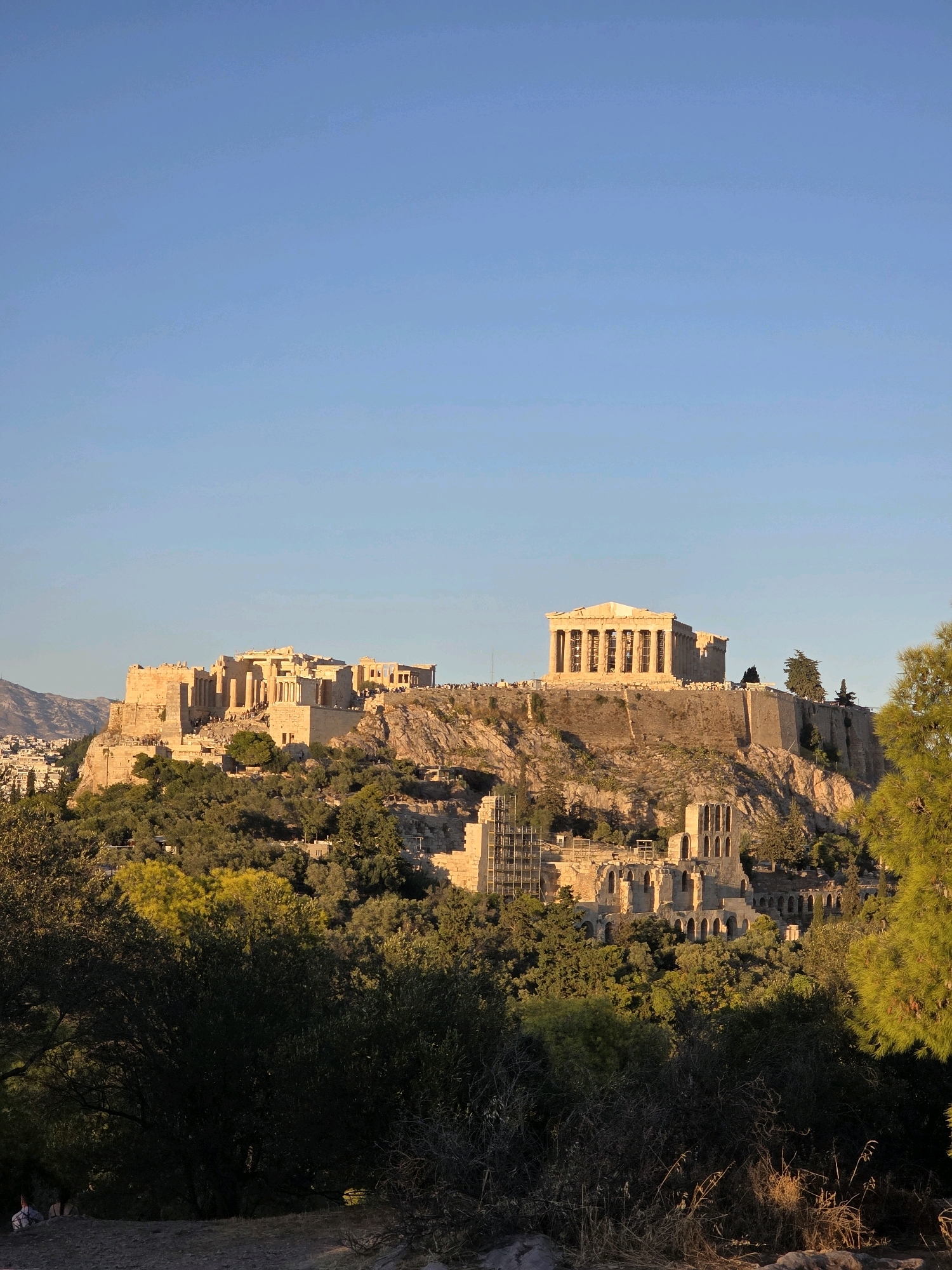 The image depicts the Acropolis of Athens, Greece, bathed in the warm light of either sunrise or sunset. The ancient citadel sits atop a rocky outcrop, overlooking the modern city. The Parthenon, a prominent temple with its iconic Doric columns, is clearly visible on the highest point of the Acropolis. Surrounding the Parthenon are other ruins and structures, some in a state of preservation and others appearing as skeletal remains. The slopes of the Acropolis are covered with lush green trees, with some foliage casting shadows onto the ancient stones. In the foreground, the lower slopes are densely wooded, with dark shadows and patches of sunlight. The sky above is a clear, bright blue, indicating good weather conditions. In the far distance, beyond the immediate structures of the Acropolis, faint outlines of hills or mountains can be seen. On the far left of the image, a few people are visible in the foreground, seated or standing amongst the vegetation, appearing to be observing the historical site. The overall impression is one of ancient grandeur set against a backdrop of natural beauty and urban sprawl.