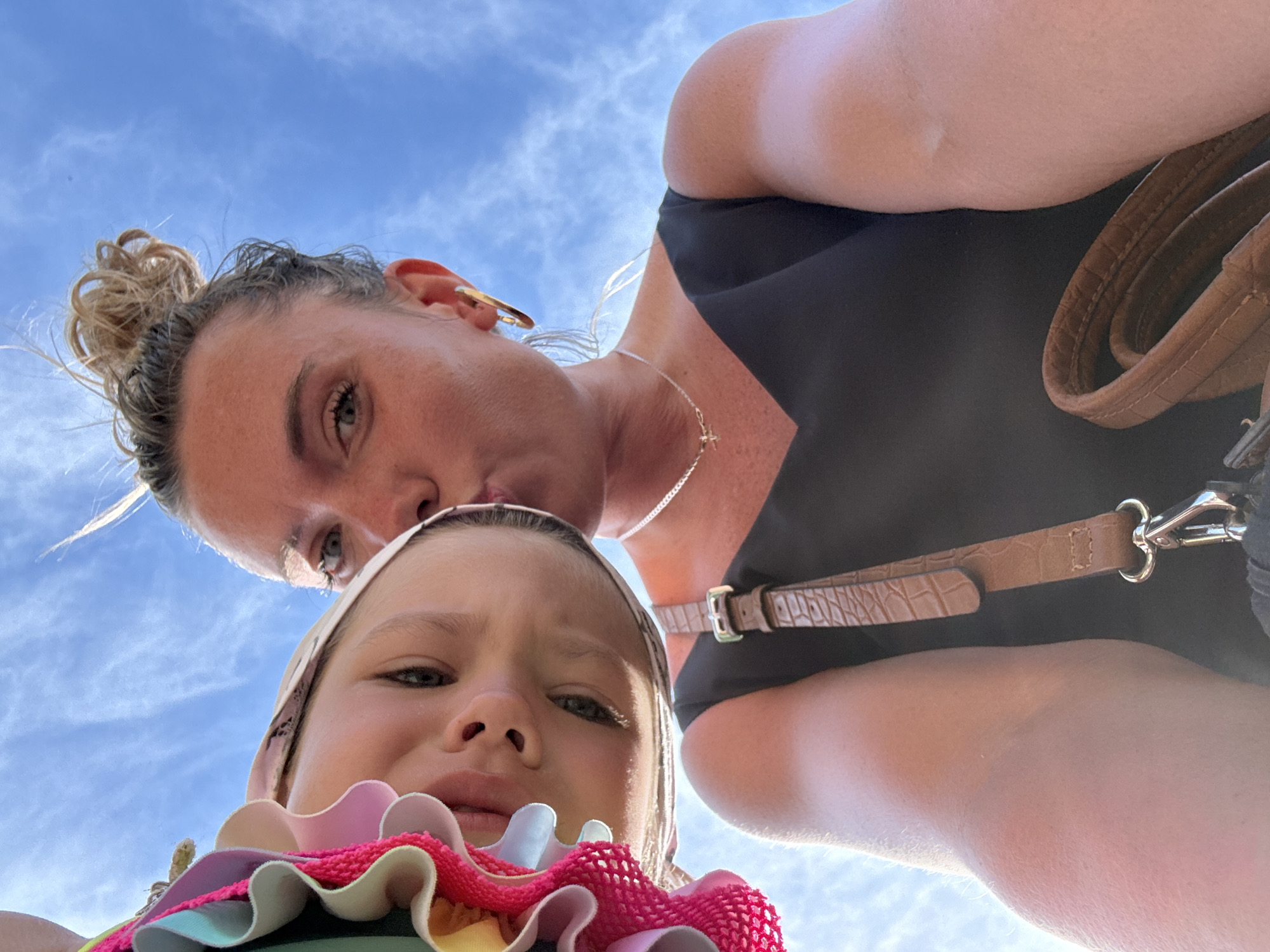 This is a selfie taken from a low angle, looking up at two people against a bright blue sky with wispy white clouds. In the foreground, a young girl with a light-colored headscarf and a colorful ruffled top is looking directly at the camera with a slightly pained or anxious expression. Her eyes are wide, and her mouth is slightly open. Behind her and slightly to the side, a woman is visible from the nose up. She has her hair pulled up in a messy bun and is wearing large hoop earrings. Her head is tilted downwards, and she appears to be making a kissing face. She is wearing a black top and a brown handbag with a textured strap is visible over her shoulder. The scene appears to be outdoors, possibly during the daytime given the bright sky. The woman is wearing a silver chain necklace with a small pendant. The setting is likely Anapa, Russia.