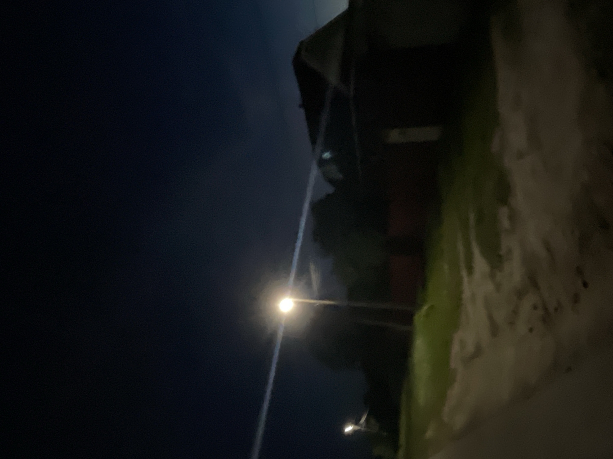 The image depicts a dark outdoor scene, likely at night, with a street lamp illuminating a dirt path and grassy areas. A house with a dark roof is visible in the background. The sky is a deep blue, suggesting nighttime. The location is stated to be Красные Баки, Russia.