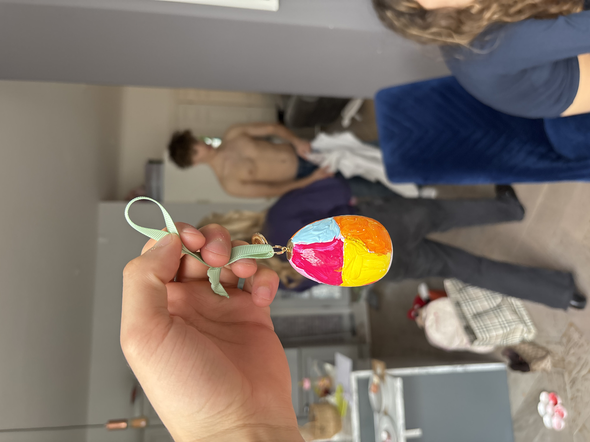 A person holds a painted Easter egg ornament with a green ribbon. The egg is divided into four sections, each painted a different color: pink, yellow, orange, and light blue. The background shows a partially visible interior setting with two other people present. One is seated on a blue upholstered item. The second person is visible from the chest up and is shirtless. The overall scene gives the impression of a casual indoor environment. There is no indication that the location is London, UK.