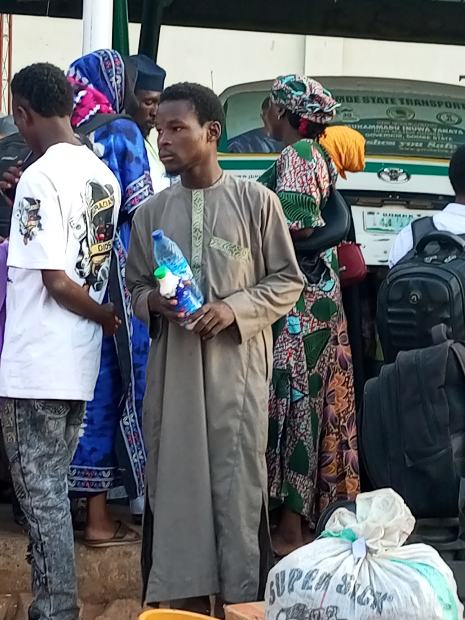 In Gombe, Nigeria, a group of people are gathered near a vehicle. The foreground features a man in a tan robe holding a blue bottle of water and a smaller white container. To his left, a person in a white t-shirt with a graphic design and distressed jeans stands. Behind them, a person in a blue and pink patterned headscarf and blue attire is visible, along with another individual wearing a black cap and a light-colored garment. To the right of the man with the water bottle, a woman in a vibrantly patterned dress and headscarf is partially seen. The background reveals the side of a white vehicle with text, including "GOMBE STATE TRANSPORT" and names that appear to be "MUHAMMADU INUWA YAYAYA" and "GOVERNOR". Several dark backpacks are also visible. The ground appears to be a mixture of dirt and stone, with a large white sack labeled "SUPER SICK" in the lower right corner.