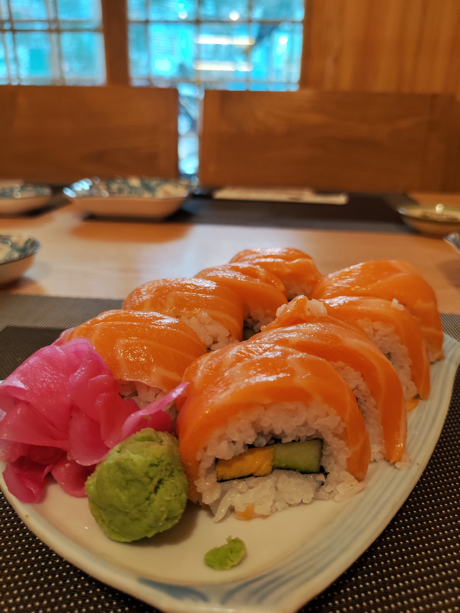 A plate of salmon sushi rolls is presented on a table in what appears to be a restaurant setting. The sushi rolls are topped with slices of salmon, with a filling that includes rice, cucumber, and possibly egg. Alongside the sushi rolls are pickled ginger and a dollop of wasabi. The plate is white with blue accents. In the background, there are other tables set with plates and bowls. The scene suggests an indoor dining environment, possibly in Da Lat, Vietnam. The presence of specific sushi ingredients and the style of plating are common in Vietnamese cuisine.