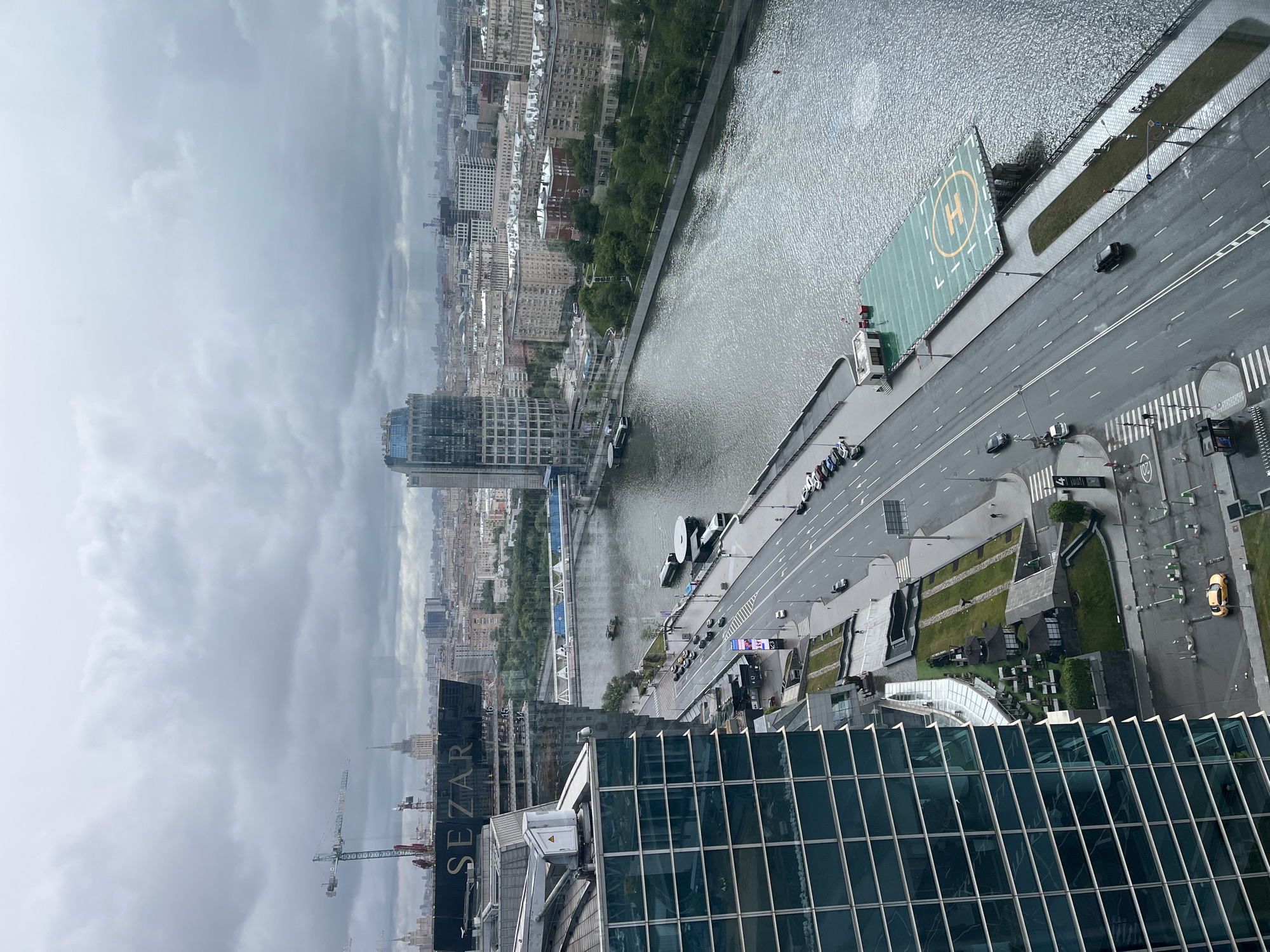 The image depicts an elevated view of an urban landscape on a cloudy day. A river winds through the city center, with numerous buildings lining its banks. A prominent helipad is visible on the river's edge. A road runs alongside the river, featuring a few cars and pedestrians. Construction cranes are present, suggesting ongoing development in the area. In the foreground, the glass facade of a tall building dominates the view. The overall scene conveys a sense of a modern, densely populated city.