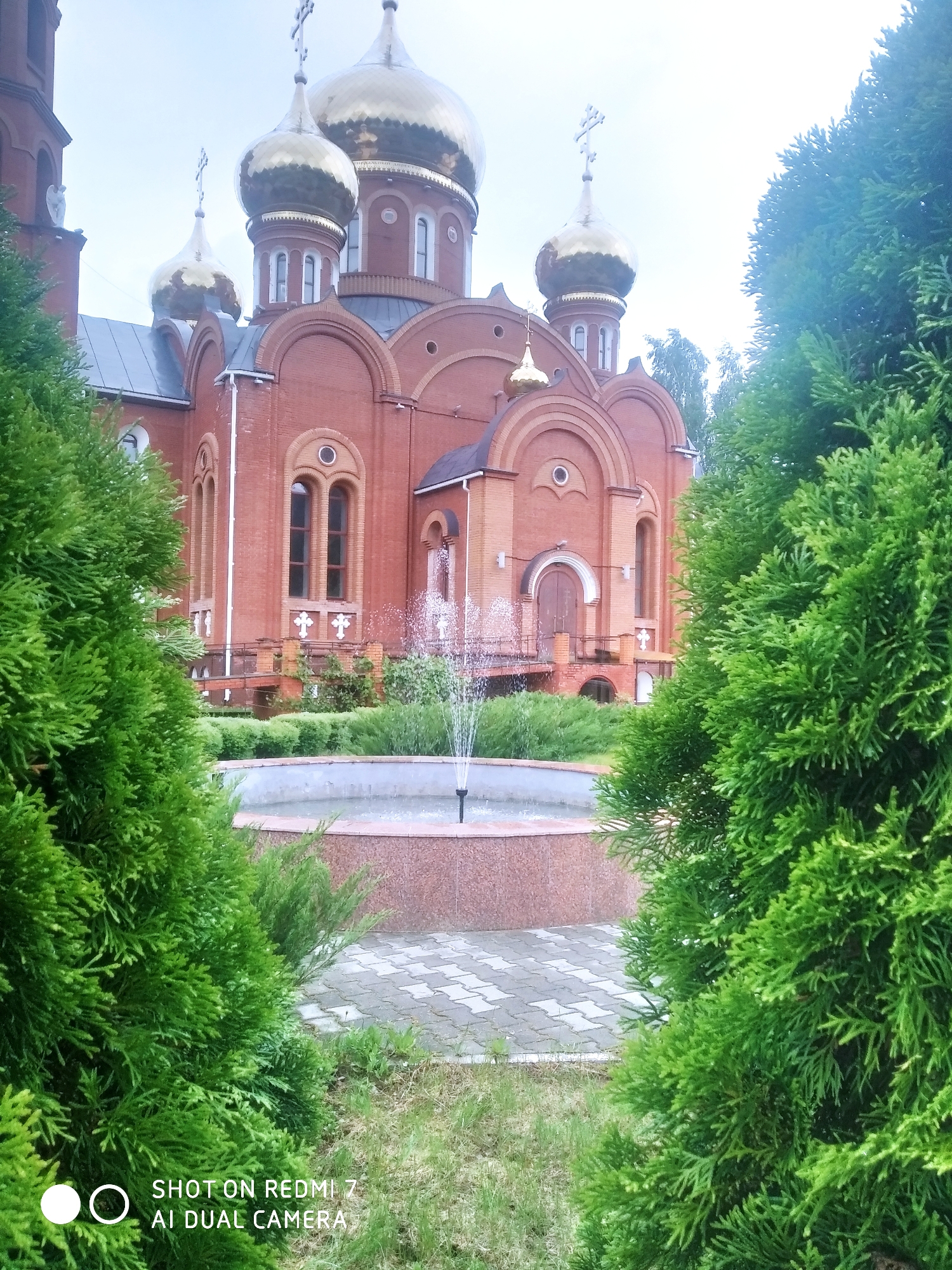The image depicts a brick building with multiple golden domes topped with Orthodox crosses, likely a church or cathedral. The structure is set back from the viewer, partially obscured by lush green coniferous trees in the foreground. A small fountain with a central jet is visible in front of the building, surrounded by low hedges. The ground is covered with gray paving stones. The sky appears overcast and gray. The text at the bottom of the image indicates it was taken with a Redmi 7 AI Dual Camera. The location is likely Chaikovsky, Russia, given the architecture of the building.