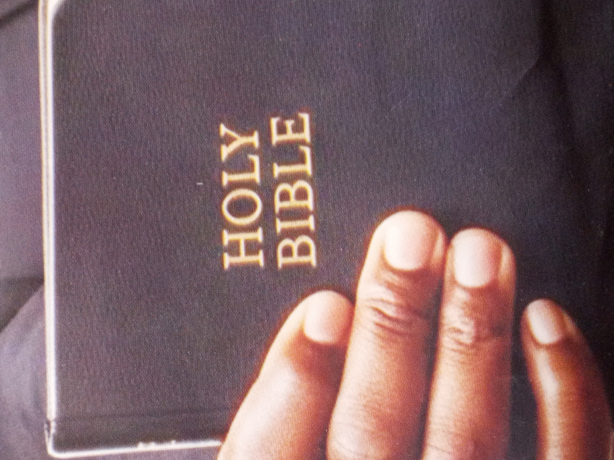 This close-up, vertically oriented image captures a person's hand resting on a dark-covered Holy Bible. The skin tone of the hand is dark, consistent with a person of African descent, situating the image in the context of Badagry, Nigeria. Four fingers are prominently visible, with neatly kept nails, and a portion of a thumb is seen on the right edge.
The book's cover is a deep, solid color, likely black or dark blue, with the words "HOLY BIBLE" embossed in golden, sans-serif capital letters across its surface. The text is oriented sideways in the frame, suggesting the book is positioned with its spine towards the top or bottom of the original shot. The edges of the book are visible on the left, showing a lighter, possibly white or cream-colored, page block or spine detail. The background is indistinct and dark, keeping the focus entirely on the hand and the sacred text. No specific activity beyond holding or resting on the book is evident, implying a moment of contemplation, reading, or devotion. The lighting is soft, highlighting the textures of the book cover and skin.