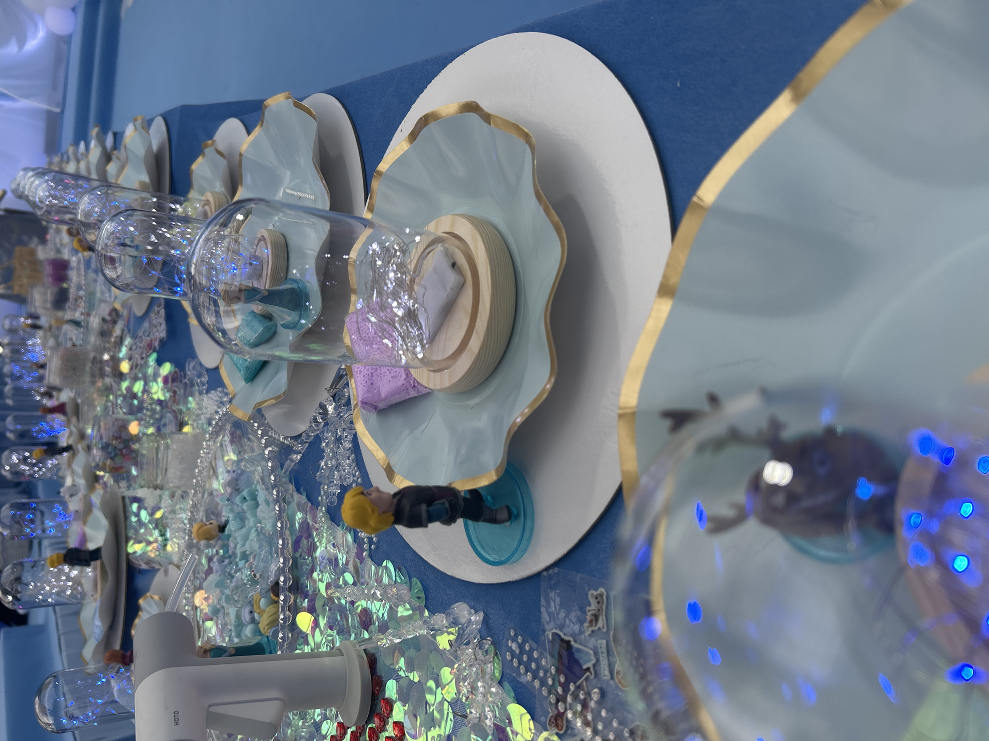This image captures a close-up view of an elaborately decorated table setting, likely for a themed children's party or event, heavily inspired by Disney's "Frozen." The scene is indoors and features a predominantly blue, white, and gold color scheme, with sparkling accents.
The table is covered with a blue cloth and set with multiple place settings. Each setting features a large white circular charger, topped with a light blue, ruffled or shell-shaped plate/bowl, intricately edged in gold. A central decoration for each setting is a clear glass dome or bottle-like container. These containers are filled with tiny blue fairy lights, creating a magical, twinkling effect, and house various small objects. One dome clearly shows a small, blonde-haired male figurine, resembling Kristoff from "Frozen," standing on a blue circular base. Another container holds a light purple, textured item, possibly a decorative sponge or toy. All these glass elements rest on light-colored wooden circular bases.
To the left of the main settings, a large section of iridescent, sequin-like material is visible, adding a shimmering, icy texture to the table decor. In the foreground, a white and gray device, possibly a dispenser, is visible, with the brand name "HOTO" clearly printed on its side. Small stickers bearing the word "FROZEN" are also visible on the blue table surface, directly affirming the party's theme. The background consists of a solid blue wall or backdrop, with some faint white circular elements.
The atmosphere suggested is festive, imaginative, and meticulously designed, aiming to create a whimsical experience for attendees. The artificial lighting inside the domes enhances the celebratory mood. No specific time of day or outdoor weather conditions are discernible, as it is an indoor event. The location context is Moscow, Russia, though no specific visual cues in the image directly indicate this.