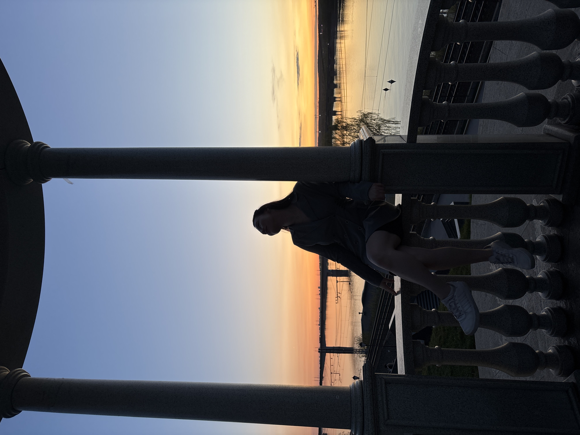 A young person sits on a stone balustrade overlooking a river at sunset in Perm, Russia. The sky transitions from blue to orange. The subject rests their legs on the lower part of the balustrade. Power lines are visible across the river.