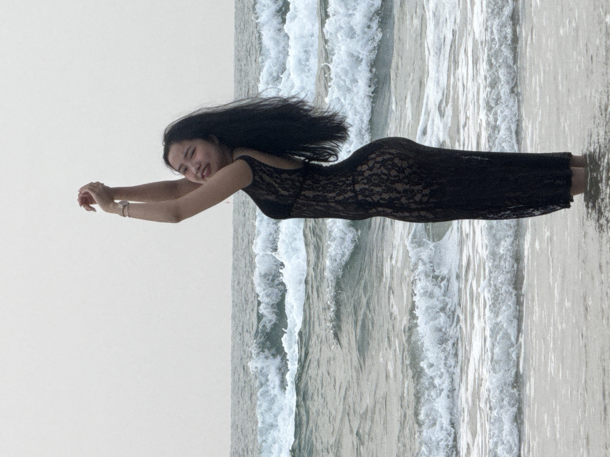 A woman in a black dress lies prone on a beach with a background of waves and overcast sky. The location is consistent with a beach in Đà Nẵng, Vietnam. The woman's arms are extended forward. She is smiling. The water is receding around her.