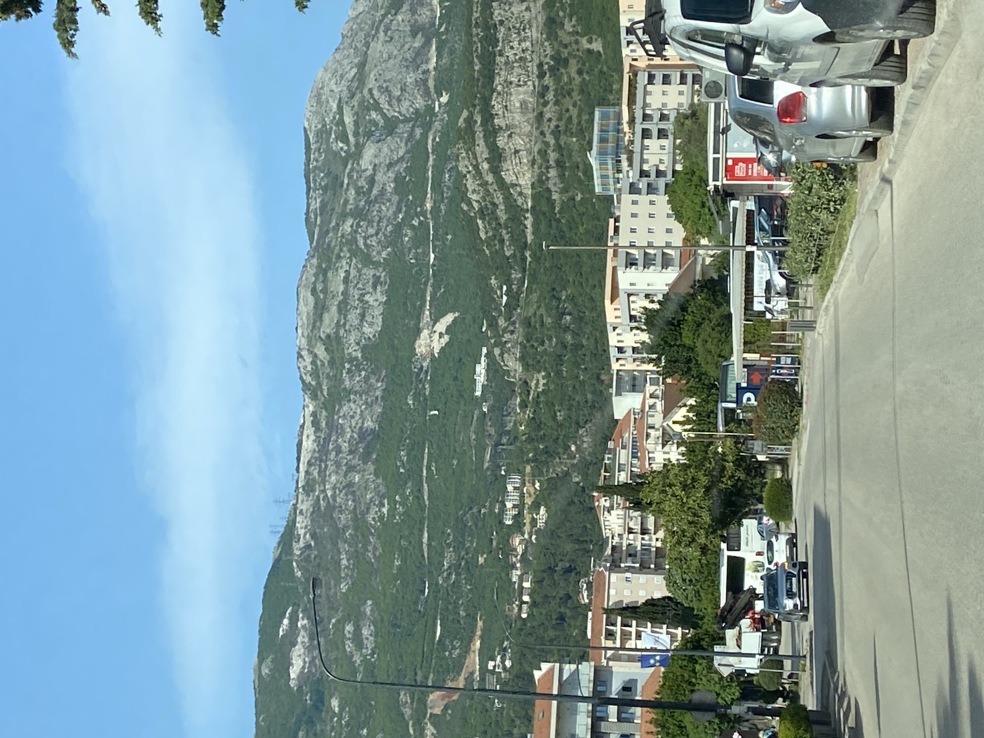 The image depicts a street scene in Budva, Montenegro, with a large mountain visible in the background. The sky is blue with minimal clouds. A street runs vertically through the right side of the image, with parked cars lining the sidewalk. Buildings of varying heights and architectural styles are situated along the right side of the street, interspersed with trees and greenery.
On the left side of the frame is a steep, rocky mountain covered in patches of vegetation. Power lines are visible along the mountainside. A street light is located near the bottom left of the image. The photo appears to be taken from a slightly elevated position.