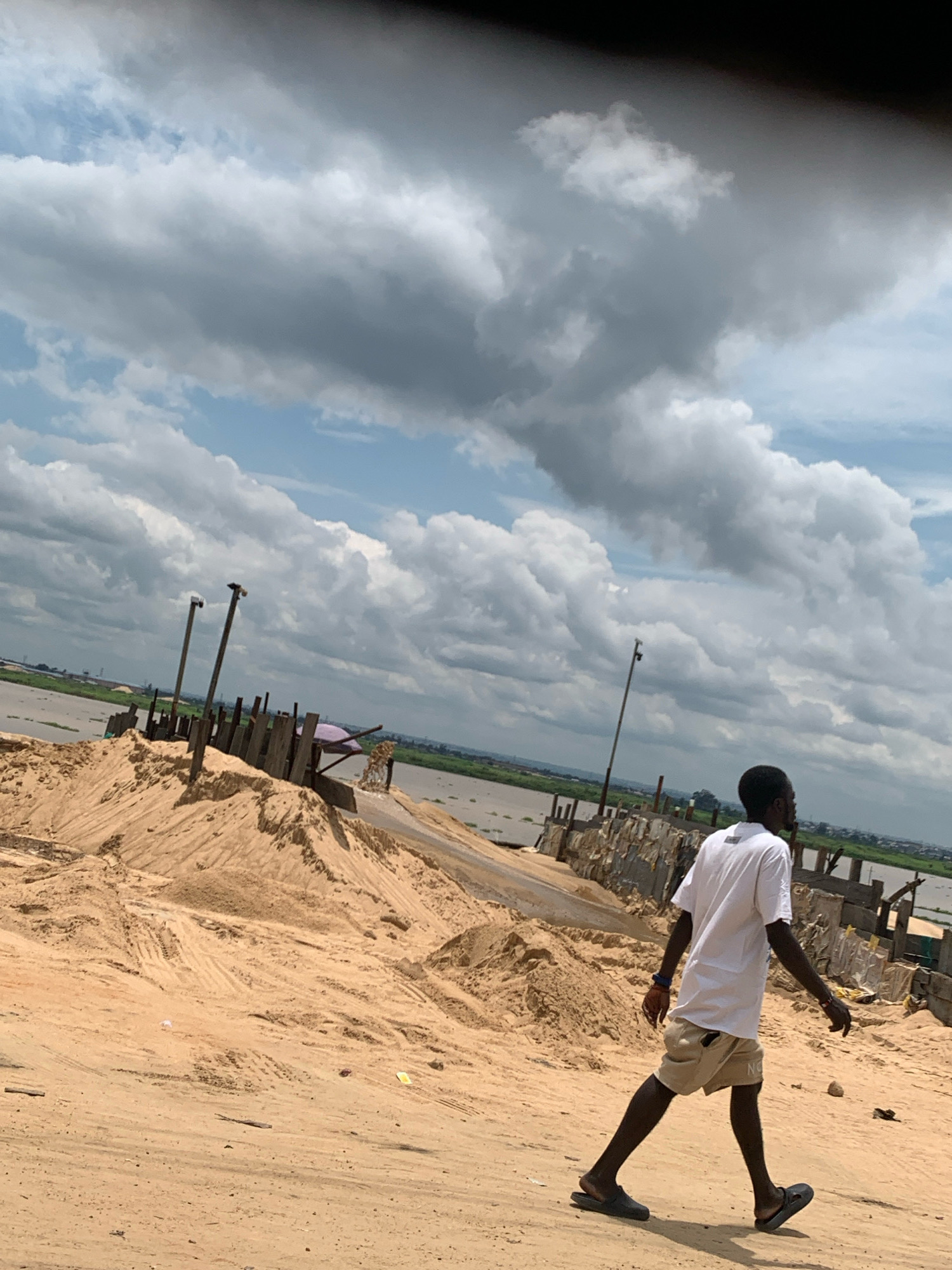 In Fegge, Nigeria, a young Black man wearing a white t-shirt and tan shorts walks across a sandy landscape. He is viewed from the side, walking from left to right, with his right leg extended forward. The environment appears to be a construction or excavation site with large mounds of sand and debris. In the background, a body of water is visible, possibly a river or lagoon, with a distant treeline and buildings suggesting a developed area. The sky is filled with dramatic, dark clouds, indicating overcast weather. Several wooden posts and some rudimentary structures are scattered across the sand.