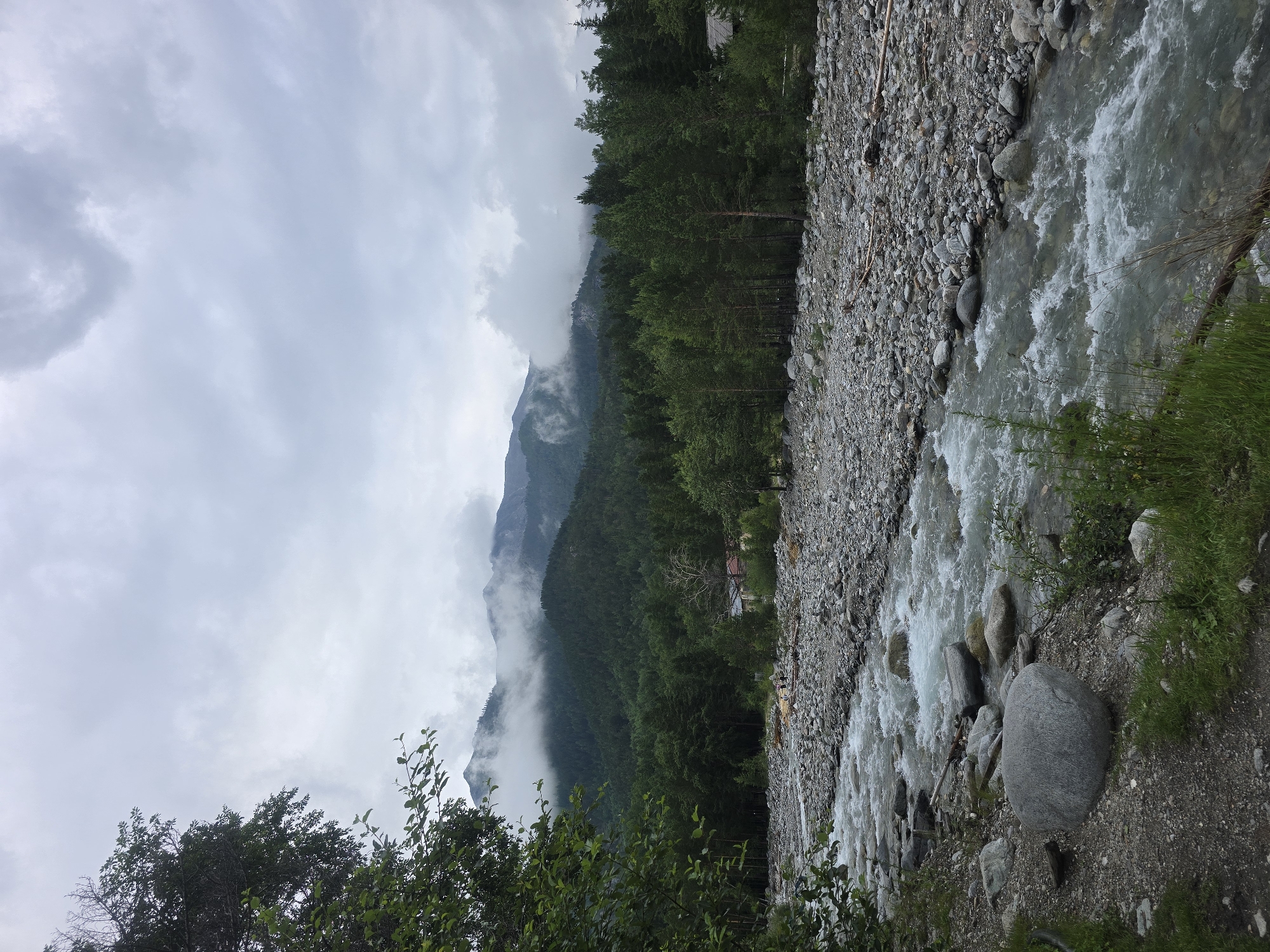 This is an eye-level, outdoor shot of a rocky riverbed in Arshan, Russia. The river flows from the bottom right of the frame towards the top left, with whitewater rapids visible. The riverbanks are lined with numerous grey and brown stones of varying sizes, interspersed with patches of green grass and small bushes. In the background, a dense forest of green pine trees covers rolling hills. The forest gives way to a mountain range in the distance, with its peaks shrouded in white clouds. The sky above is overcast and a pale grey. A few small, rustic buildings with red roofs are nestled among the trees in the mid-ground. Several people are visible on the riverbank in the distance, some appearing to stand on a log bridge.
