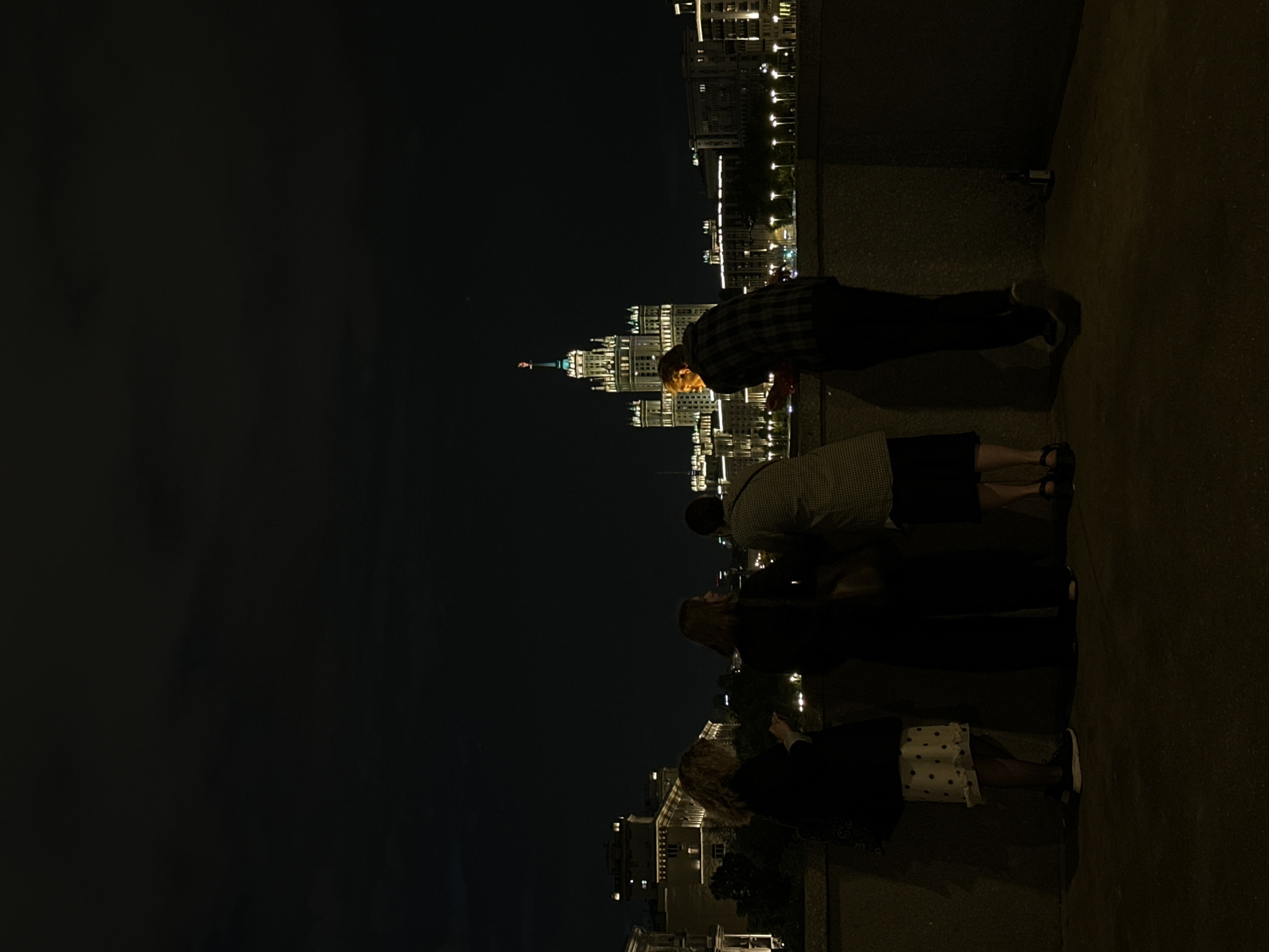 A nighttime scene shows a group of at least four people looking out over a parapet with the lights of Moscow, Russia in the background. The top of a Stalinist skyscraper is prominently featured, likely one of the Seven Sisters. People stand on a pavement or walkway in front of the parapet. The overall lighting is dim, with the city lights and building illumination providing contrast.