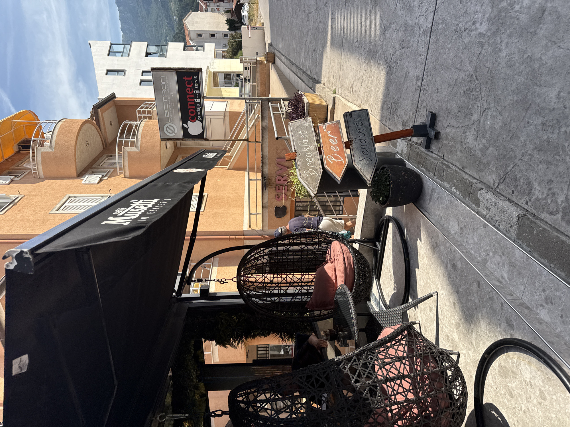 This is a street-level, medium-shot photograph taken in the city of Budva, Montenegro, on a sunny day. In the foreground, on the left, are two hanging wicker chairs with pink cushions. A small black table is visible between them. To the right, a signpost displays three directional signs: one pointing left reads "Breakfast," another pointing right reads "Beer," and the bottom sign pointing left reads "Sendviči." A potted plant sits at the base of the signpost, and another plant is visible behind the chairs.
Behind the chairs, a woman wearing a blue shirt and a blue baseball cap walks away from the camera towards a building with an orange facade. The building has several balconies with white railings. A black awning is stretched over the seating area.
Above the orange building, a white building stands, and further in the background, mountains are visible under a partly cloudy sky. A street or sidewalk extends into the distance on the right side of the frame. A white car is parked on the right side of the street in the background. The overall scene suggests an outdoor seating area for a cafe or restaurant.