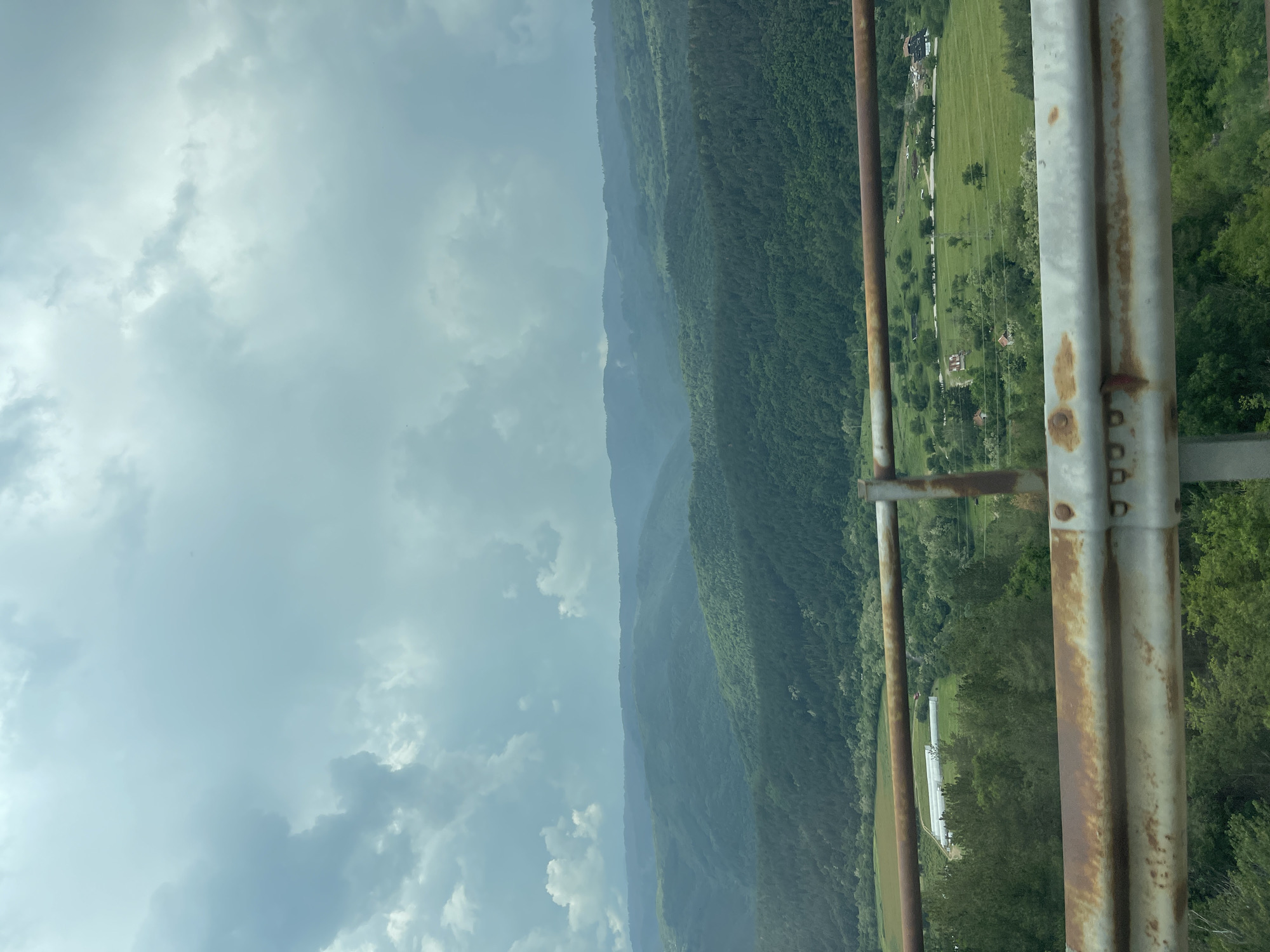 The image presents a wide, elevated view of a rural, mountainous landscape under an overcast sky. In the immediate foreground, a two-tiered, rusted metal guardrail runs horizontally across the lower portion of the frame, indicating the vantage point is from a bridge or an elevated road. Beyond the guardrail, a valley floor spreads out, characterized by vibrant green fields and numerous deciduous trees.
In the midground, several elongated, white or light-colored structures, resembling agricultural greenhouses or tunnels, are visible within a field on the left side of the valley. Further to the right in the midground, scattered residential or farm buildings with visible roofs are interspersed among fields and groves of trees. A power line structure or pole is also present in this section. The predominant color of the vegetation is various shades of green, indicative of lush foliage.
The background is dominated by multiple successive layers of heavily forested mountains and hills, stretching into the distance. These mountains appear progressively lighter and hazier towards the horizon, a visual effect of atmospheric perspective. The sky above is entirely covered by a thick layer of grey and light blue-grey clouds, with some areas appearing brighter than others, suggesting a generally diffused light without direct sunlight. No individuals or specific human activities are discernable within the scene. This landscape is located in the region of Klisura, Bulgaria.