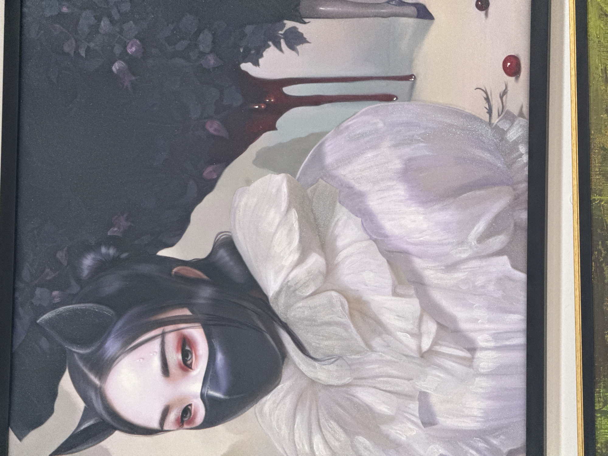 The image is a framed painting, possibly on canvas, depicting a figure in a dark, surreal setting. The subject is a pale-skinned person with black hair styled with cat-like ears. They wear a black face mask that covers their nose and mouth. Their eyes are visible and appear to have red eye shadow. The person is wearing a white, ruffled garment.
Behind the figure is a dark, blurry background of what appears to be foliage with some purplish elements. Above the figure, there's a white area with two streaks of red fluid (possibly blood). There are also two red berries near the top right corner.
The painting is framed in black, with white and gold trim visible on the right side. The photograph appears to be taken indoors, possibly in a gallery or home setting. There's no information in the image that suggests it is located in Izhevsk, Russia.