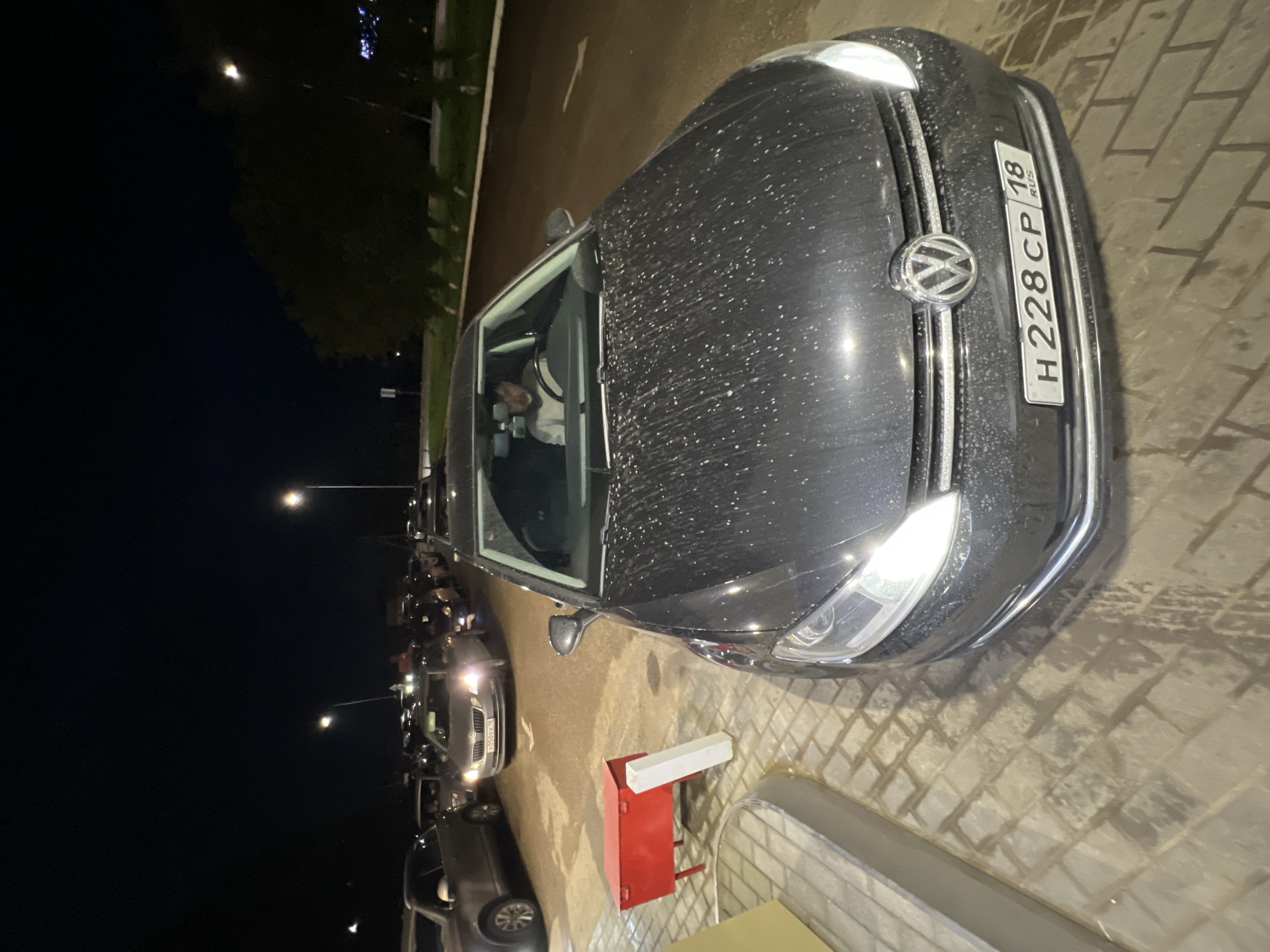 The image shows a dark colored Volkswagen parked at night in what appears to be a parking lot or street in Agryz, Russia. The car's license plate is visible and reads "H 228 CP 18 RUS". The car is facing forward, and the driver's side is slightly angled towards the camera. A person can be seen in the driver's seat. The car is covered in what looks like water droplets or dust. The ground is paved with bricks. In the background, other cars are parked in a line, and streetlights illuminate the scene. The overall lighting suggests it is nighttime, and the presence of water droplets on the car could indicate recent rain or dew. The setting appears to be an urban or suburban area.