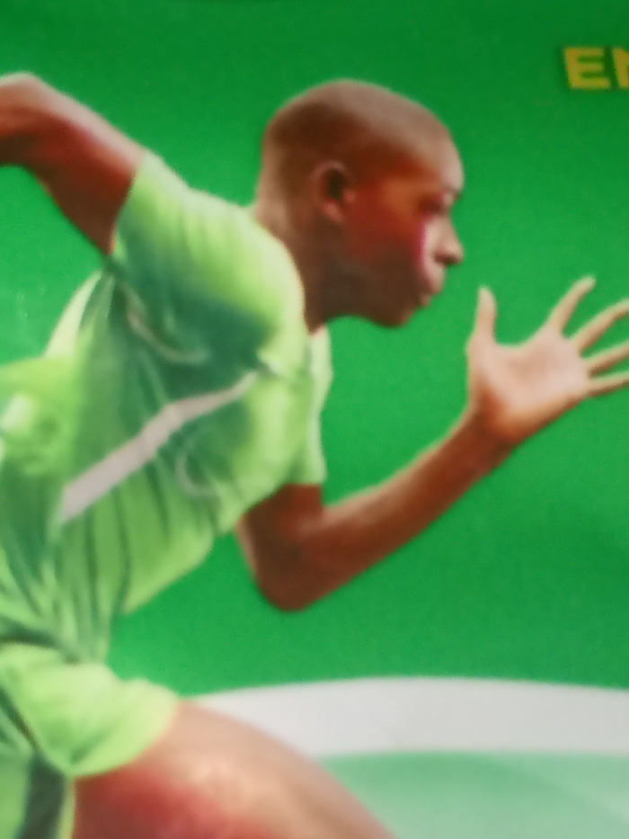 This image, captured in Adankolo, Nigeria, displays a dark-skinned individual, likely male, engaged in a dynamic running or sprinting motion. The subject is wearing a bright green, short-sleeved athletic shirt that features a white diagonal stripe or pattern across the torso. The individual's right arm is bent at the elbow and extended forward, with the hand open and fingers splayed. The left arm is bent and positioned rearward. The head is depicted in profile, facing forward. The background is a uniform bright green, with a distinct horizontal white line visible across the lower section of the frame. In the upper right corner, partial yellow letters, specifically "E" and "N", are discernible against the green background. The image exhibits significant motion blur, impacting overall clarity.
