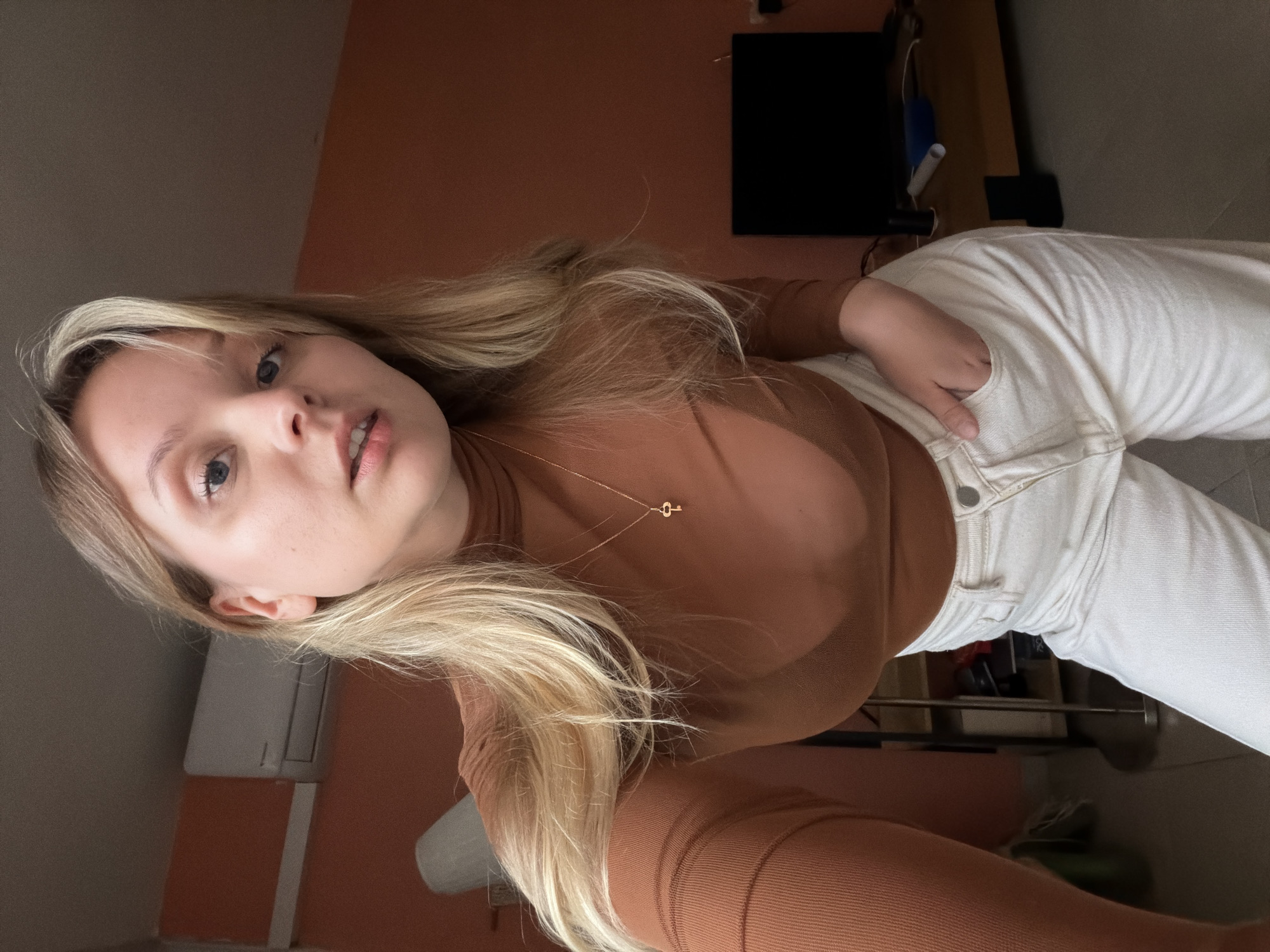 A woman with blonde hair is posing for a selfie. She is wearing a brown turtleneck shirt and white jeans. She has a gold necklace with a key pendant. The background includes an orange wall, a black television, and a white air conditioning unit. The location is likely indoors in Tel Aviv, Israel, based on the presence of the air conditioning unit, a common feature in that city.