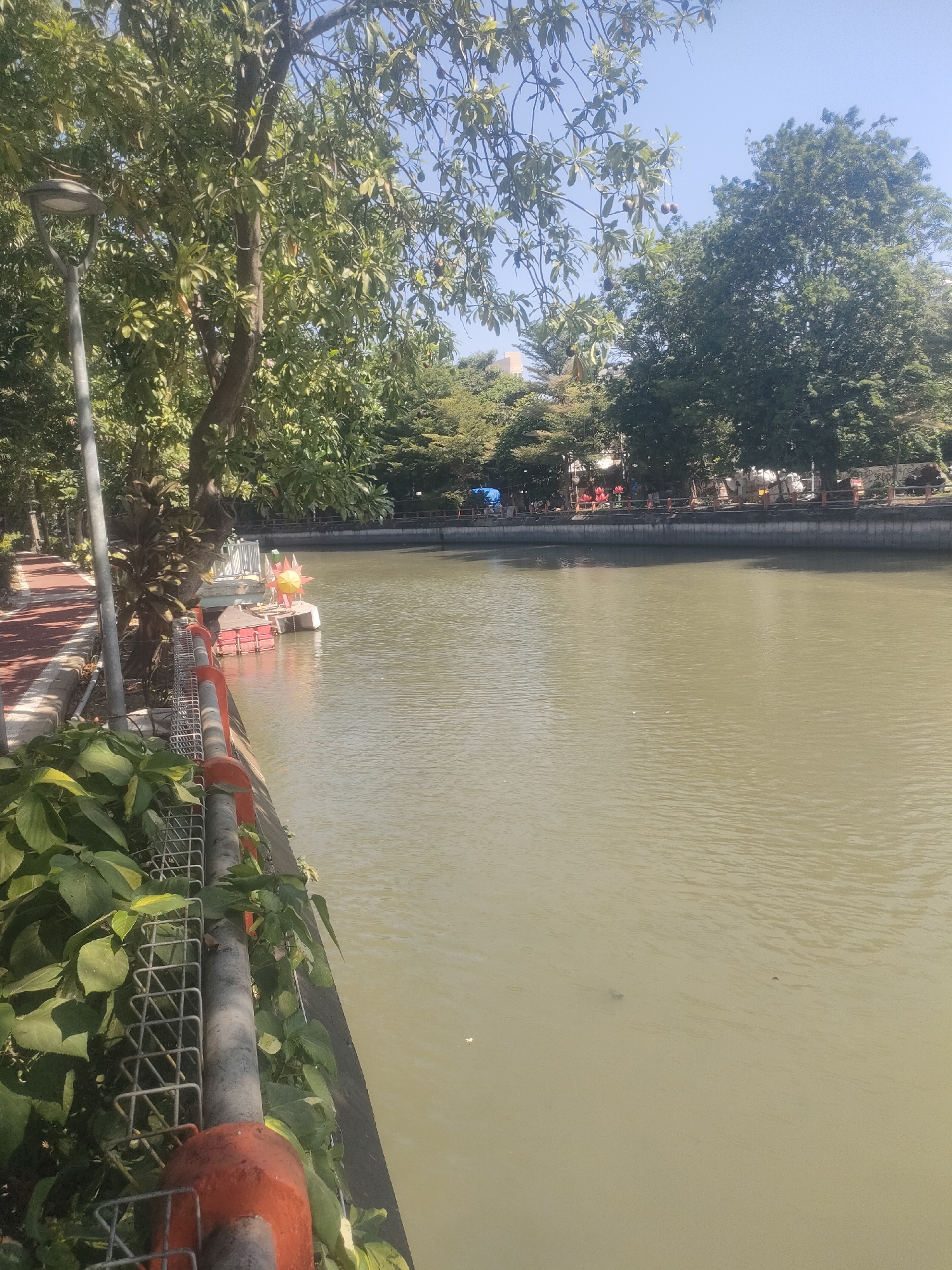 The image depicts a scenic view of the Kalimas River in Surabaya, Indonesia, from a promenade. The promenade, paved with red tiles, is bordered by lush green foliage and a metal railing with orange accents. A street lamp stands tall on the left side of the promenade.
The river, a murky green, occupies the majority of the frame. In the distance, the opposite bank is lined with trees and a low concrete wall. A few structures are visible behind the trees, suggesting urban development. A small, colorful boat, adorned with a yellow, sun-like structure and red components, is moored on the river near the promenade.
The sky is a clear, bright blue. The overall atmosphere is tranquil and inviting, likely a popular spot for recreation and relaxation within the city.