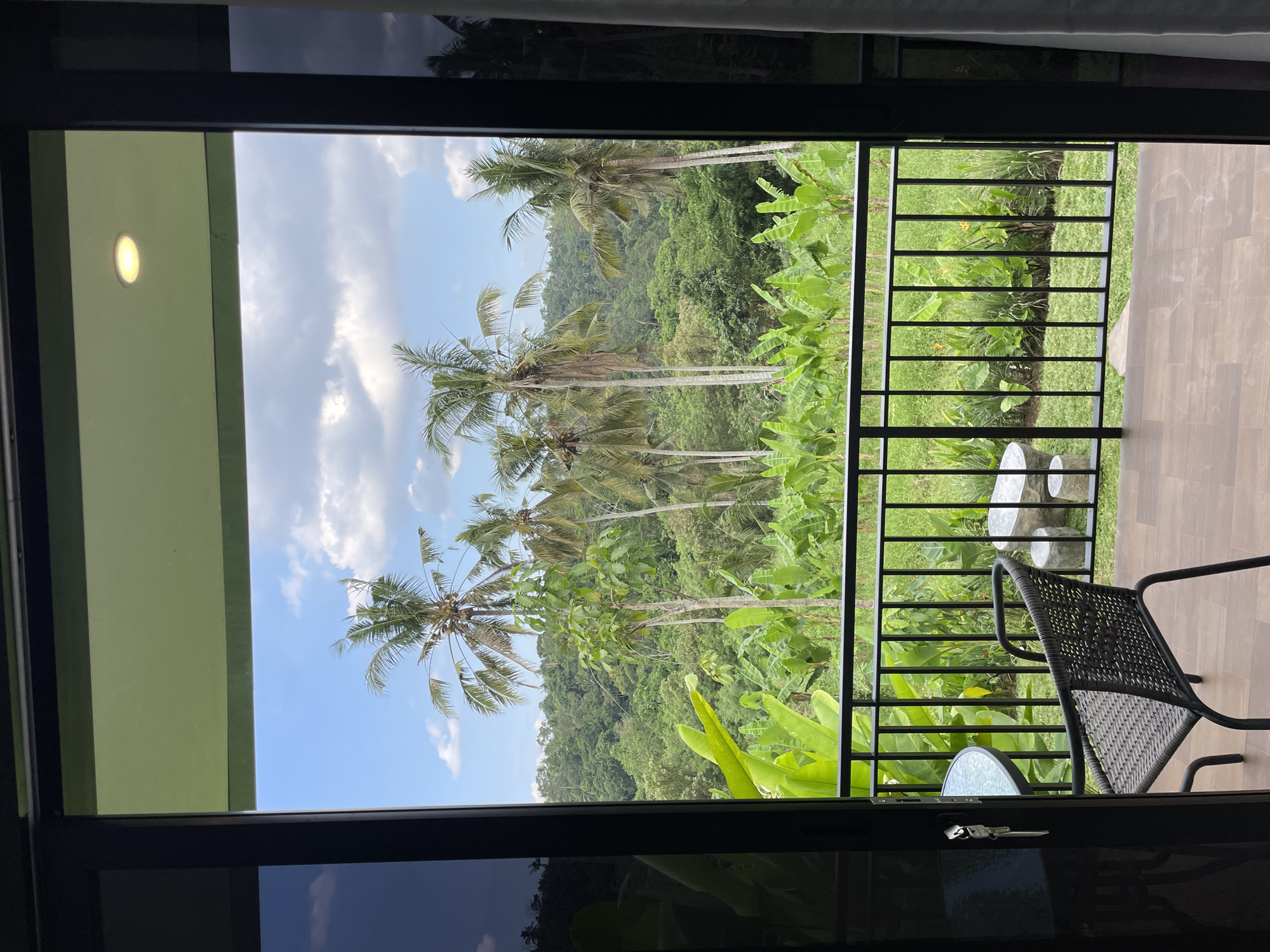 The image shows a view from a hotel room's balcony in Penebel, Indonesia, looking out over a lush, tropical landscape. Tall palm trees and dense green foliage dominate the scene, with a vibrant, clear blue sky dotted with white clouds overhead. In the foreground, a black metal railing encloses the balcony, which is furnished with a woven chair, a glass-topped table with keys resting on it, and two stone seats. The balcony floor is made of brown wooden planks. The setting appears to be a tranquil, natural environment, characteristic of rural or resort areas in Indonesia.