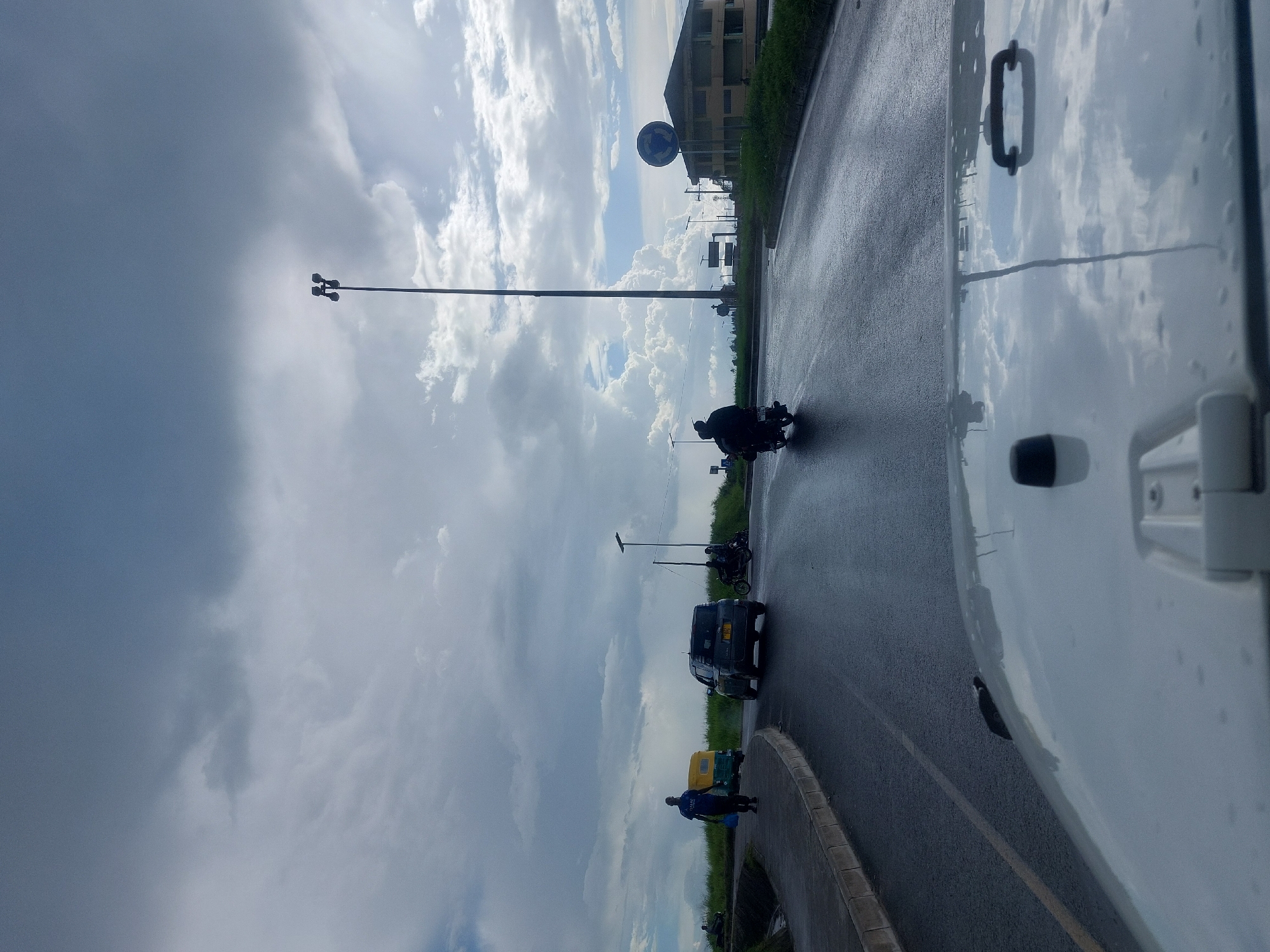 The image shows a street scene in Dar es-Salaam, Tanzania on a cloudy day. The street is wet, suggesting recent rain. A motorcyclist is riding on the road. Other vehicles visible include a dark-colored car and a yellow auto-rickshaw (tuk-tuk). A pedestrian is walking along the side of the road. The perspective is from within a vehicle, possibly a car or truck, as part of the hood is visible. Streetlights line the road. A building can be seen in the background on the right side of the image.