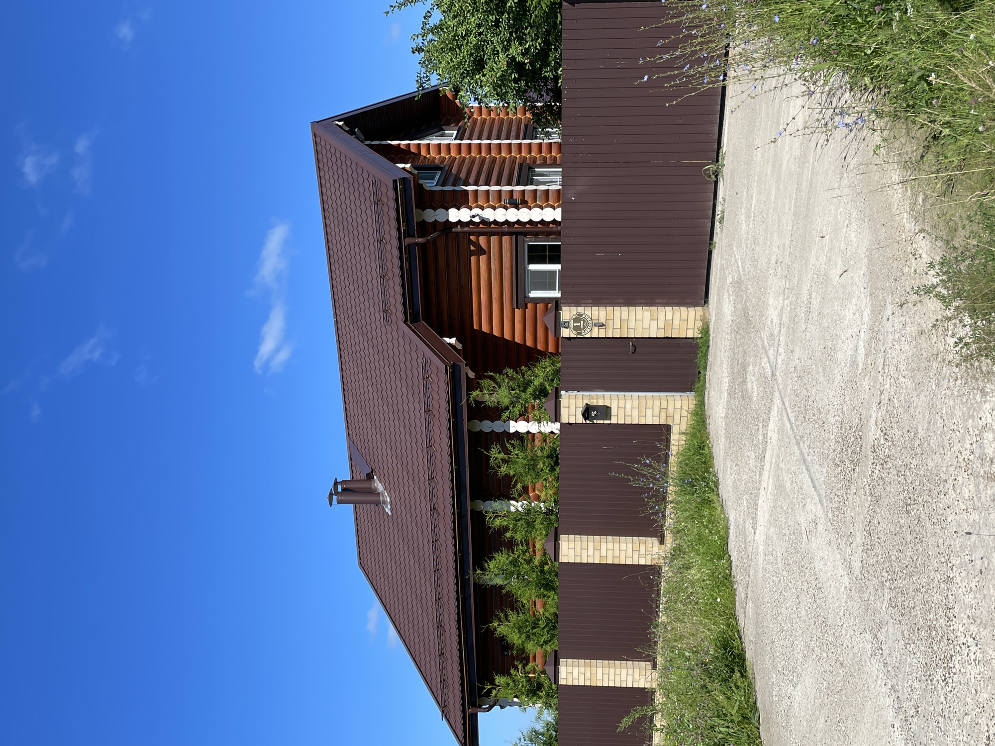 A log cabin-style house with a brown metal roof and brown siding is situated behind a brown metal fence with tan brick pillars. The house has white trim around the windows and roof edges. A brown chimney protrudes from the roof. Evergreen trees are planted in a row along the fence in front of the house. A concrete driveway leads up to the fence and a metal gate. The sky is bright blue with a few white clouds. The surrounding area is grassy with some wildflowers.