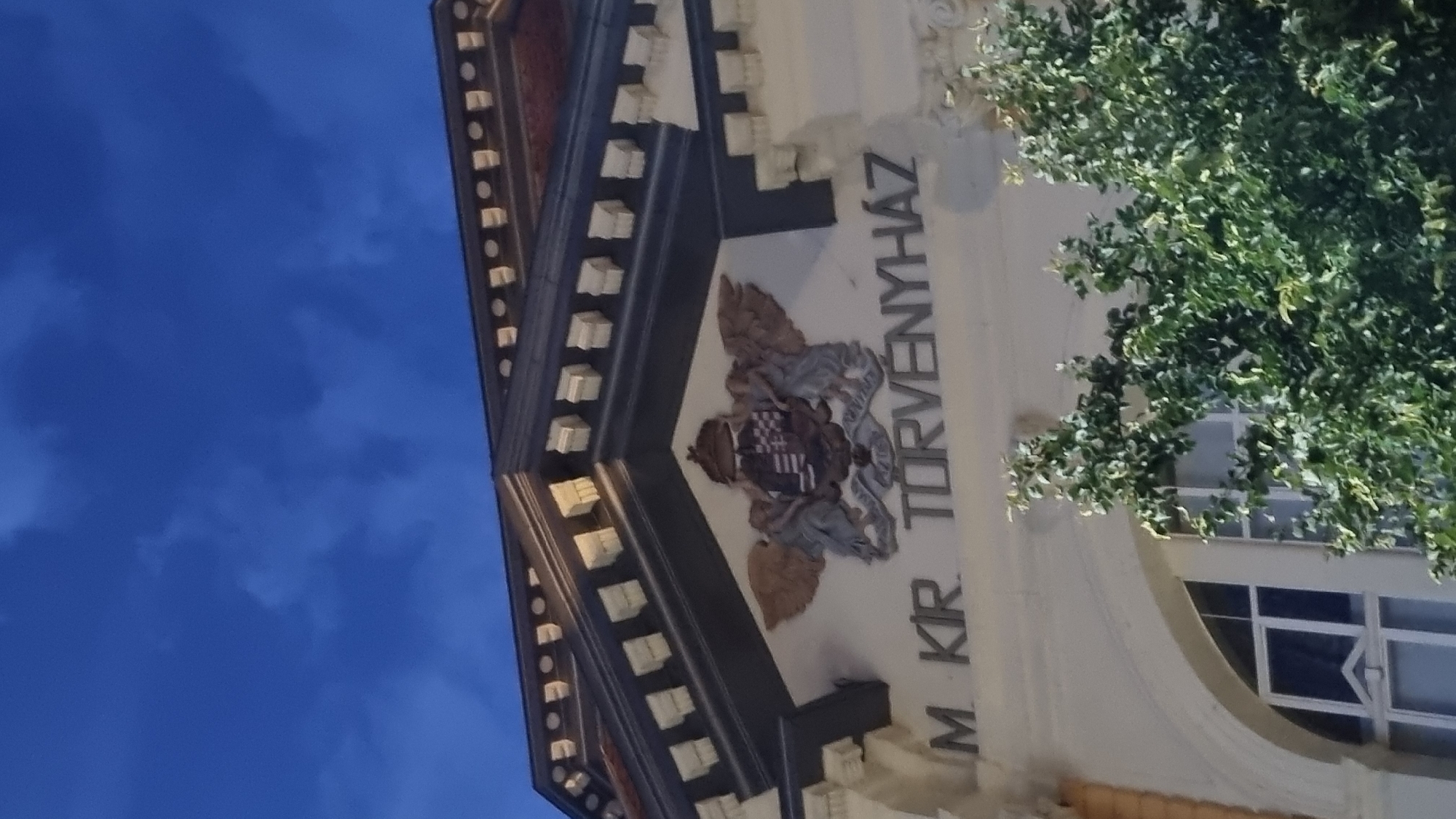 The image captures a detail of a building's facade in Berehove, Ukraine, at dusk. The sky above is a deep, cloudy blue. The building features ornate architectural elements, including decorative eaves with repeating square blocks and a prominent crest above a sign. The crest is a heraldic shield with a crown, supported by winged figures and a banner beneath. The sign, written in block letters, reads "M. KIR. TORVENYHAZ". Lush green foliage from trees partially obscures the lower portion of the building, including an arched window with multiple panes. The overall impression is of a historical or governmental building.