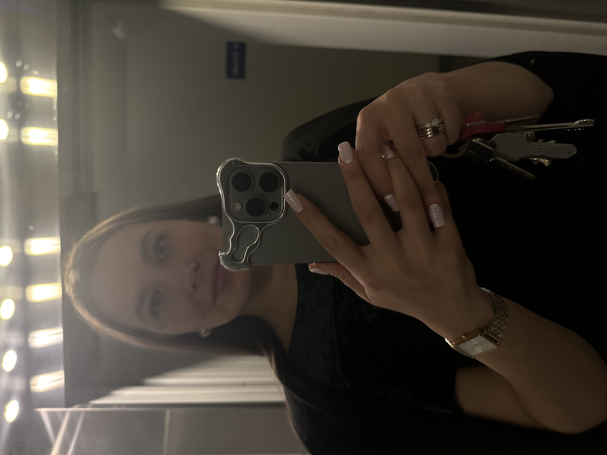 A person takes a selfie in a mirror. The background appears to be an interior space with a vertical light fixture on the left. The person is holding a smartphone with a clear case. They are also holding a set of keys. The person is wearing a watch and rings. A sign is visible on the wall behind them. The location is Odintsovo, Russia.
