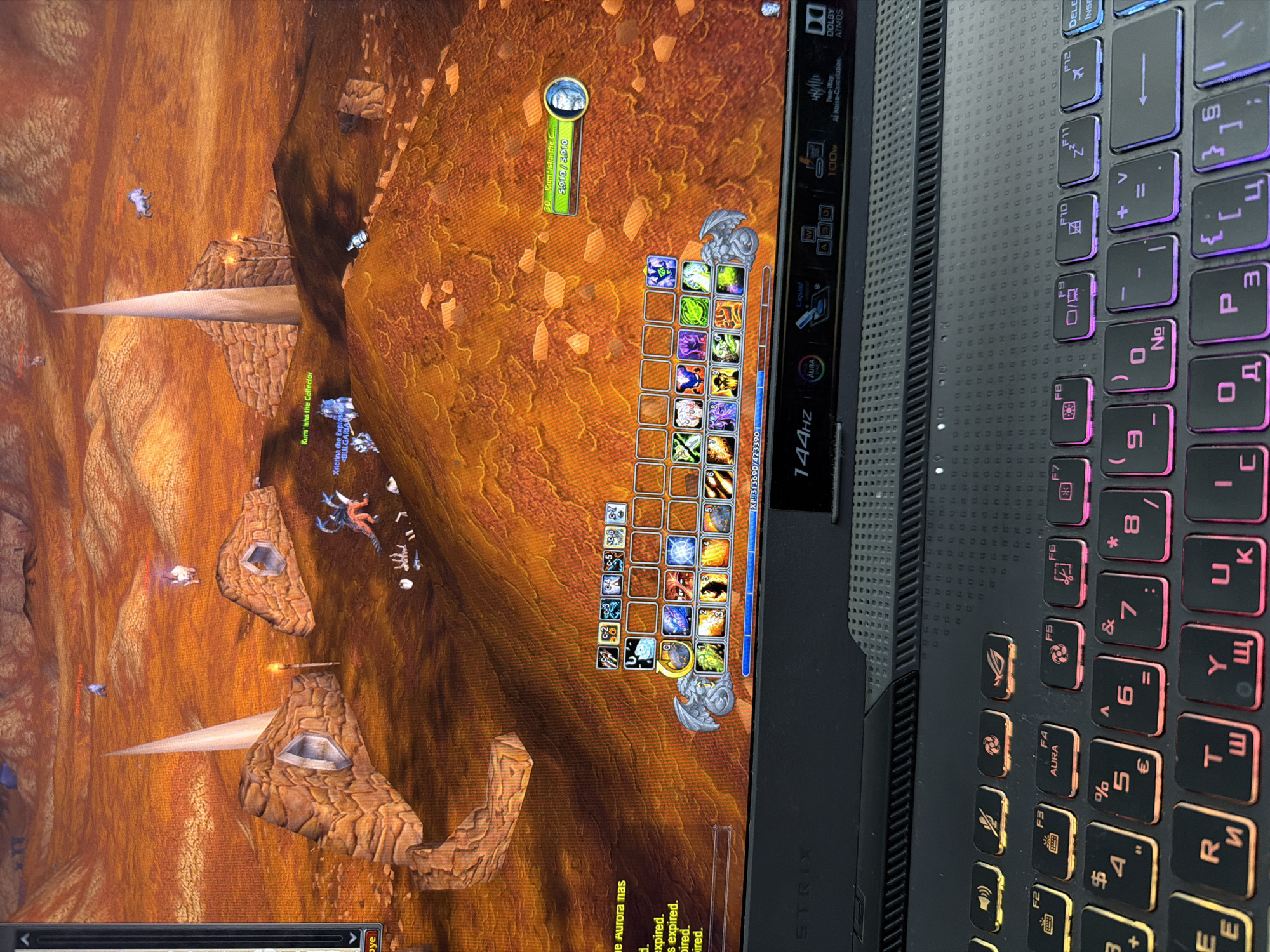 The image depicts a scene from the video game World of Warcraft being played on a laptop. The game's graphical interface is visible, showing a character in a desolate, rocky, orange-hued landscape with scattered structures and distant mountains. A character portrait and various action bars with icons are displayed on the screen. Below the game screen, the keyboard of a black ASUS ROG laptop is visible, with some keys illuminated by customizable RGB lighting. The laptop also has several dedicated function buttons above the keyboard, including one labeled "144Hz." There is no indication that this scene is located in the city of Plovdiv, Bulgaria.