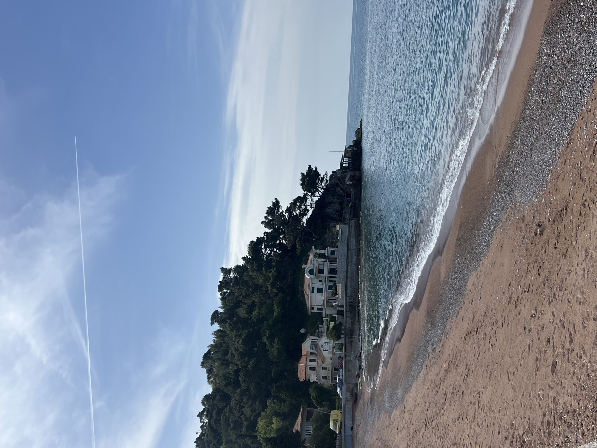 The image depicts a beach scene in Petrovac centar, Montenegro. The view is from the shoreline, looking out at the sea and the coastline. A sandy beach stretches along the foreground, with visible footprints in the sand. The sea is light blue, with waves gently washing onto the shore. Along the coast, there is a dense green forest, with several buildings nestled within the trees and along the waterfront. The sky is mostly clear blue, with a few streaks of clouds and a contrail from an airplane. The image is rotated 90 degrees counter-clockwise.