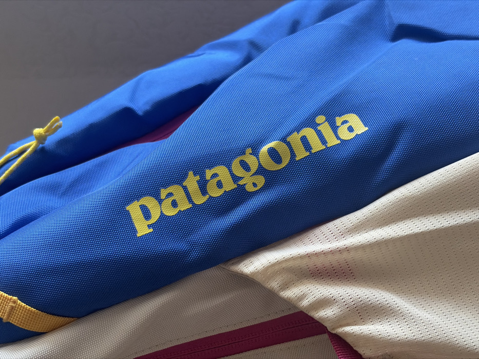 A close-up, angled shot displays sections of multi-colored technical apparel. The primary subject is a vibrant blue fabric, characteristic of an outdoor jacket or similar garment, with the brand name "patagonia" printed vertically in bright yellow, lowercase letters on its surface. To the immediate left, a section of cream-colored mesh fabric is visible, separated from the blue material by a thin, vertical magenta strip. A yellow fabric strap or trim is present near the top left edge, attaching to the blue garment, while a yellow knotted drawstring is observed on the blue fabric in the upper right. The blue material exhibits a structured, finely textured weave, and the cream fabric features a distinct mesh pattern. The background is an indistinct, plain light grey surface, suggesting an interior setting. No individuals or specific actions are depicted. The specific location in the city of Krasnaya Zarya, Russia, cannot be determined from the visual evidence provided in the image.
