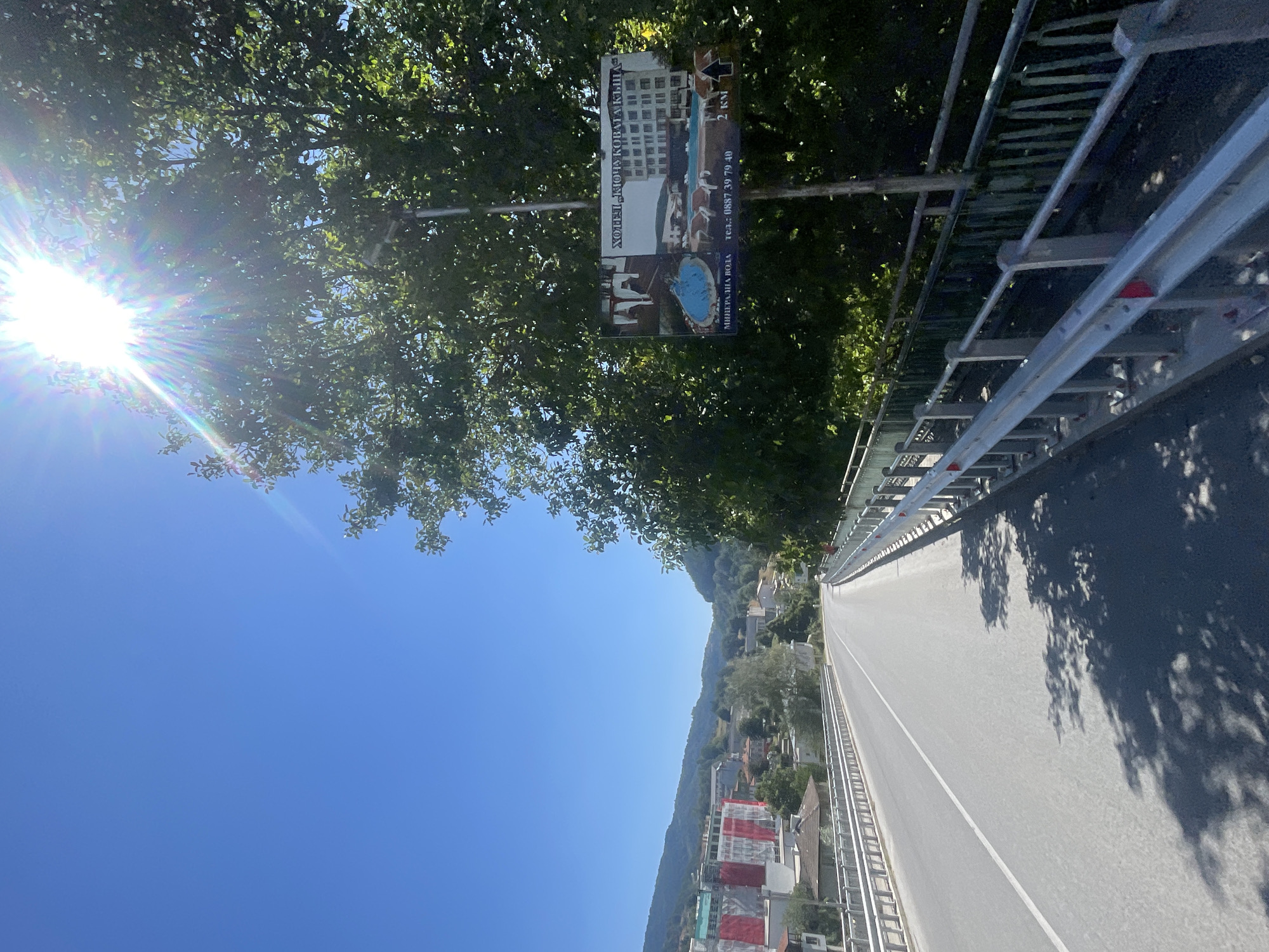 The image captures a sun-drenched scene on a road, likely in or near the city of Banite, Bulgaria. The sun is intensely bright, with visible lens flare and rays shining through the leaves of a large tree on the right side of the frame. The road, paved with asphalt, curves gently to the right, and has a white lane marking. Guardrails line both sides of the road.
In the background, a town or village is visible nestled amongst rolling, green hills. Buildings with red and white facades are prominent, suggesting construction or renovation. Further in the distance, more houses and trees dot the landscape under a clear, bright blue sky.
A signpost stands near the tree on the right. The sign itself is a billboard advertising a hotel, "HOTEL KRYCHOVATA KLYUCHKA". The advertisement features images of a swimming pool and a building, along with text that appears to be in Cyrillic. Below these images, text reads "MINERALNA VODA TEL: 0887 39 79 40" and "2 KM", indicating proximity to a mineral water source and the hotel. The sign is positioned to be visible to travelers on the road. The overall atmosphere is one of a bright, sunny day in a scenic, possibly rural or semi-rural Bulgarian setting.